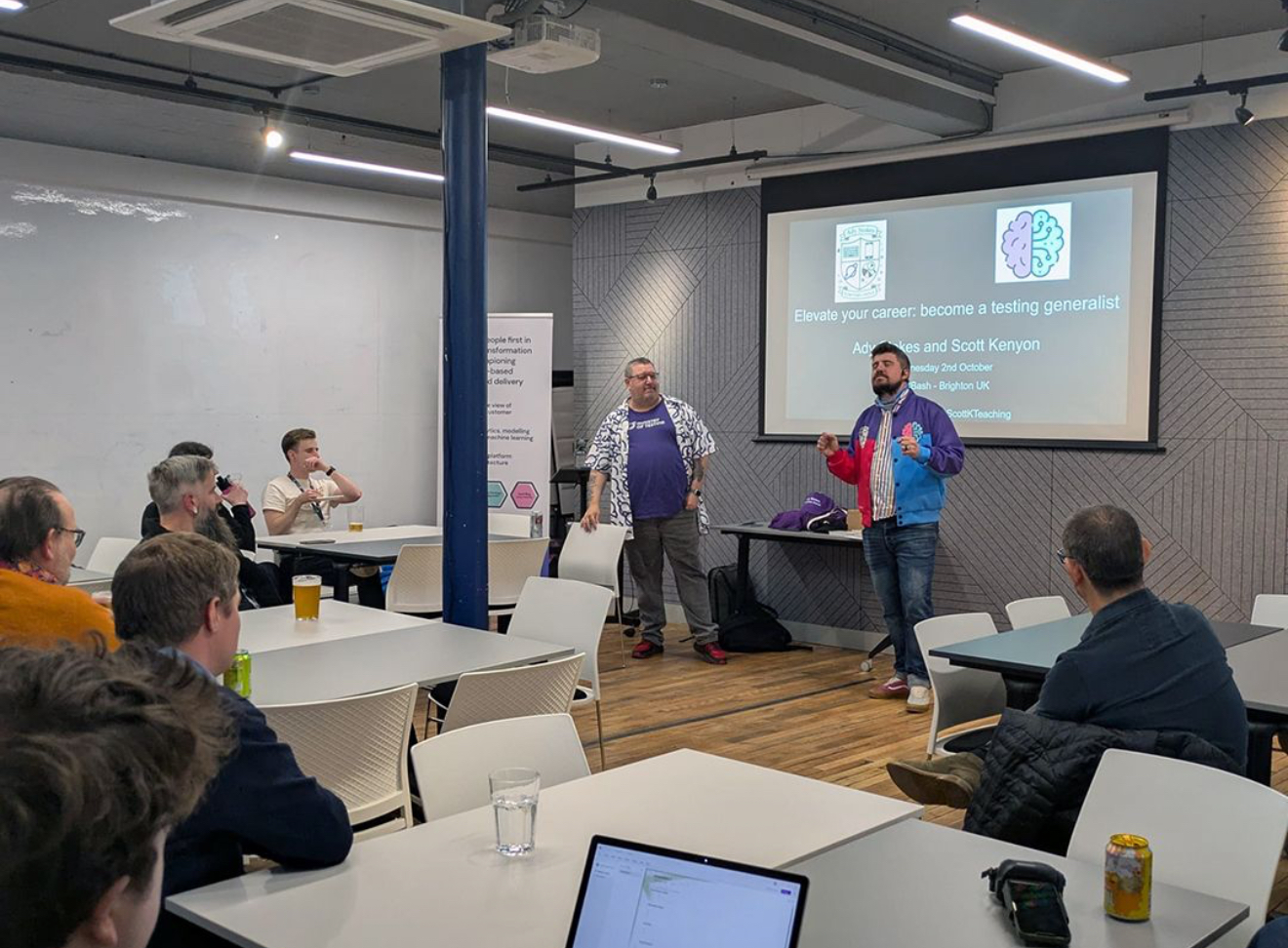 Ady and Scott stand in front of a screen with the session title how to elevate your career; become a testing generalist. Scott is wearing a pink and blue jacket with purple shoulders and Ady has a MoT planet logo Hawaiian style shirt. A group sat at several tables are looking at them. 