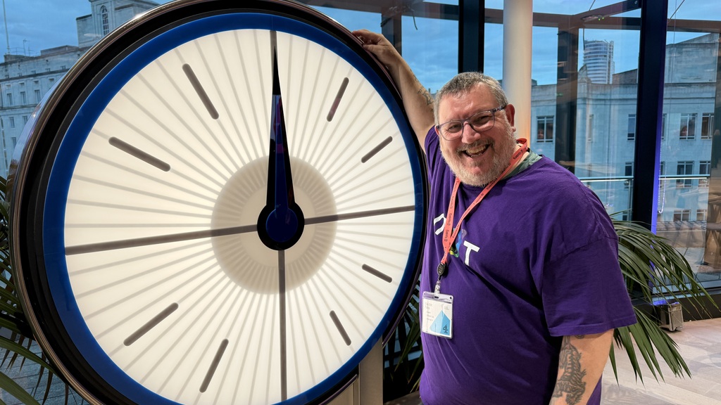 Ady in a MoT purple T-shirt with a rainbow logo is stood in front of the channel 4 countdown clock from the TV quiz.  image