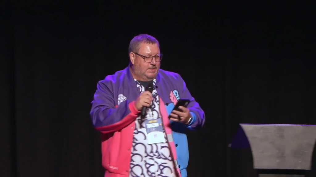 Ady Stokes on stage at MoTaCon. Microphone in one hand, phone in the other, and the captions say, "Hello. I'm Eddie S... image