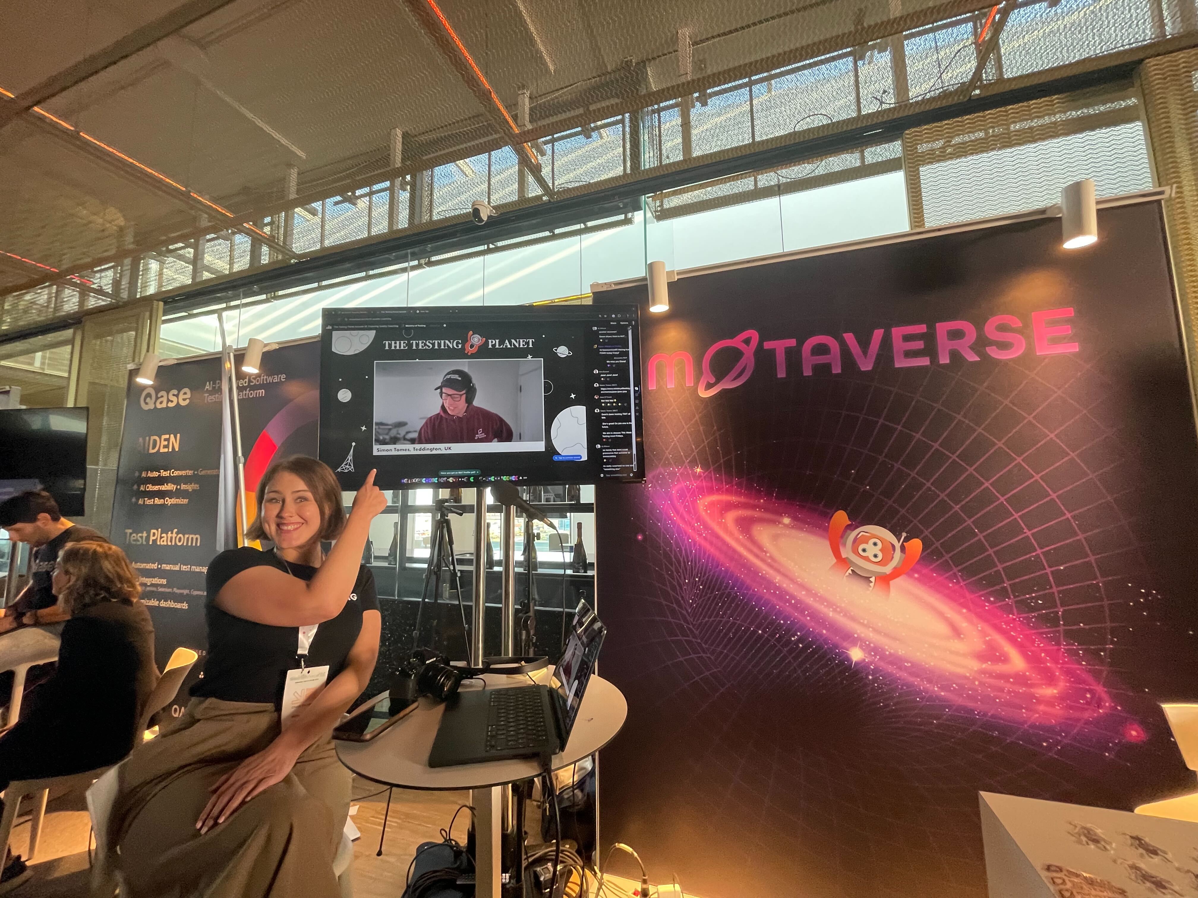 Alexandra sits in a chair pointing at a TV screen which features Simon Tomes at The Testing Planet.