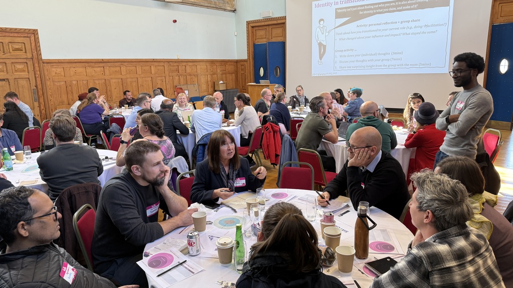 A diverse group of adults is seated at round tables in a large, bright room with wood-paneled walls and tall, leaded-glass windows, engaging in a discussion or workshop. On a projector screen at the front, a slide titled "Identity in transition" is visible, showing bullet points about "Self-reflection" and "Generating new ideas". In the foreground, one table is the focus, where several people are looking at each other and talking, with coffee cups, bottles, and papers scattered across the white tablecloth. A man in a light grey shirt stands to the right of this table, speaking or listening intently to the group. The atmosphere appears to be one of collaborative learning and networking.