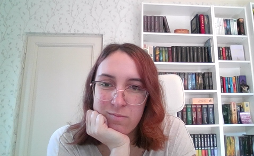 Young woman with red hair and glasses, head resting on ehr hand. There is a door and bookcase int he background.