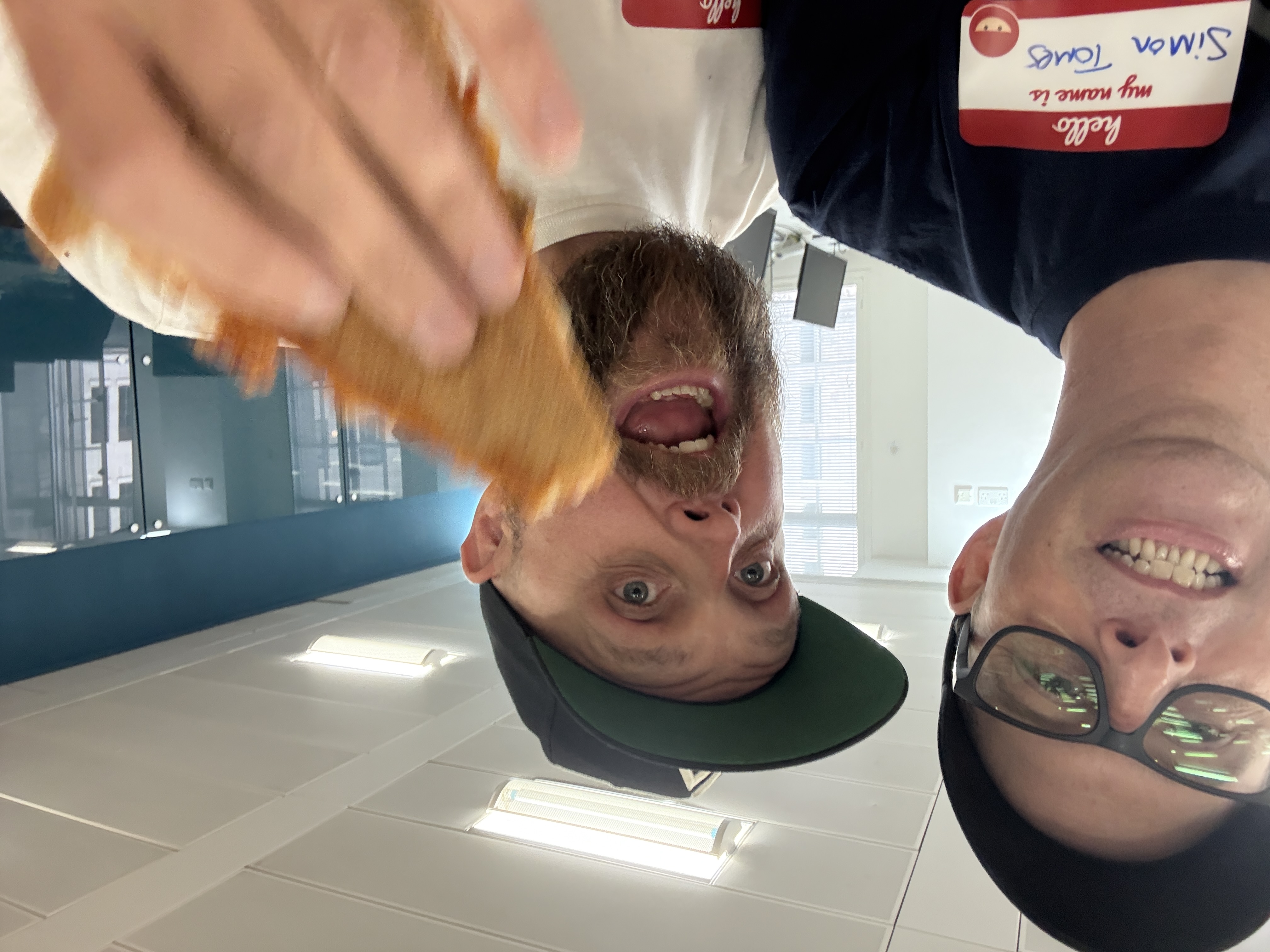 Two men with caps pull silly faces for a close-up selfie. One of them is holding up a blurred pizza slice. 