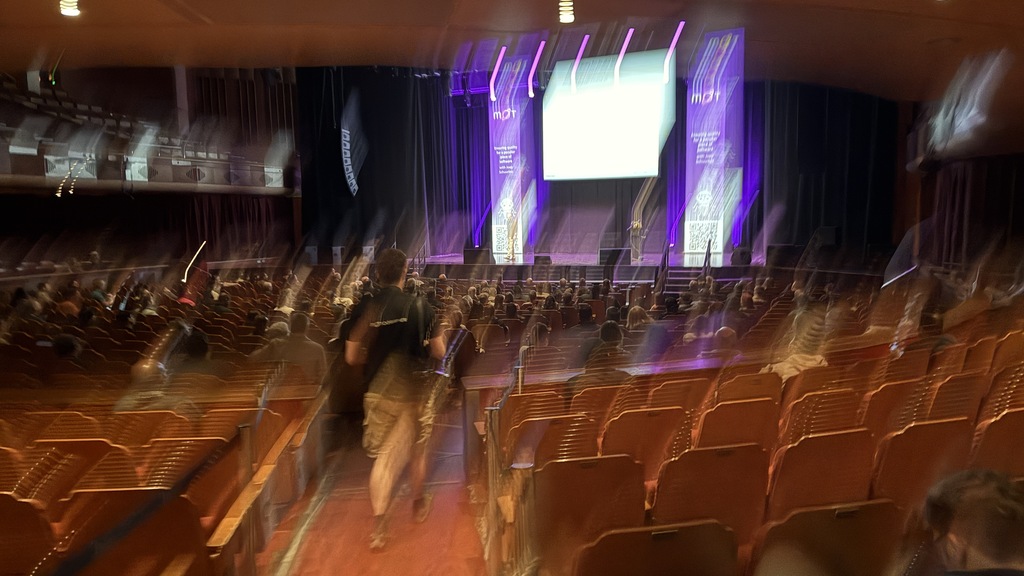 A long exposure, motion-blurred image of a large auditorium from the back. On the stage, a large bright screen is centred between two banners with a purple design and the letters 'MOT' visible. A few people are walking down the central aisle toward the stage, and the motion blur emphasises their movement.