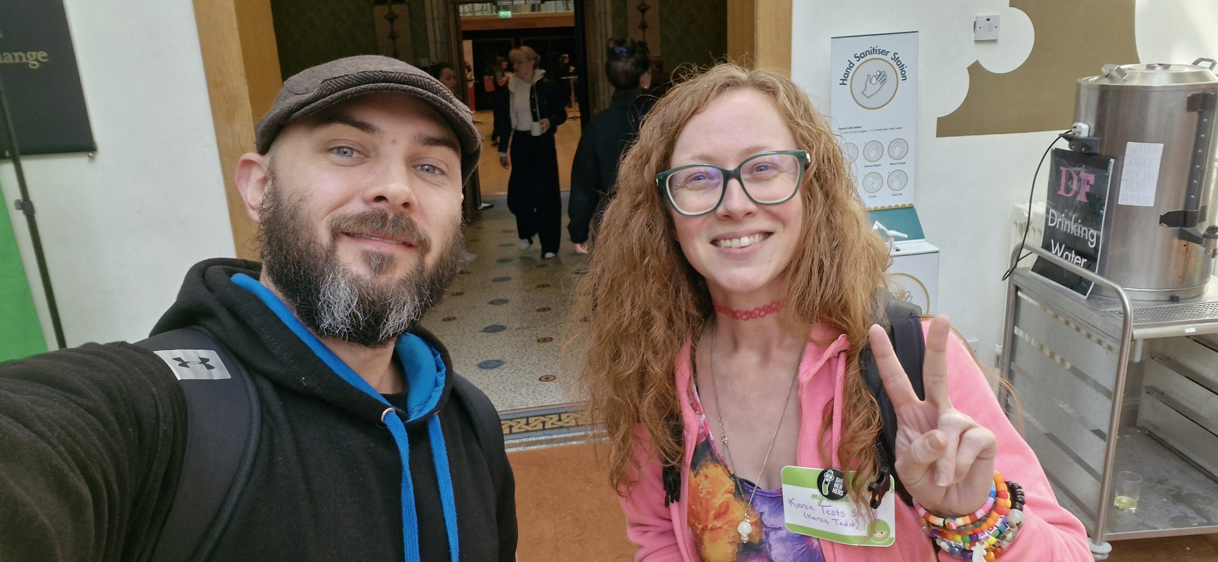 A bearded man in a cap and a woman with glasses and long red hair take a selfie indoors. The woman wears a pink jacket and a name tag while flashing a peace sign. People are visible in the background through an open doorway.