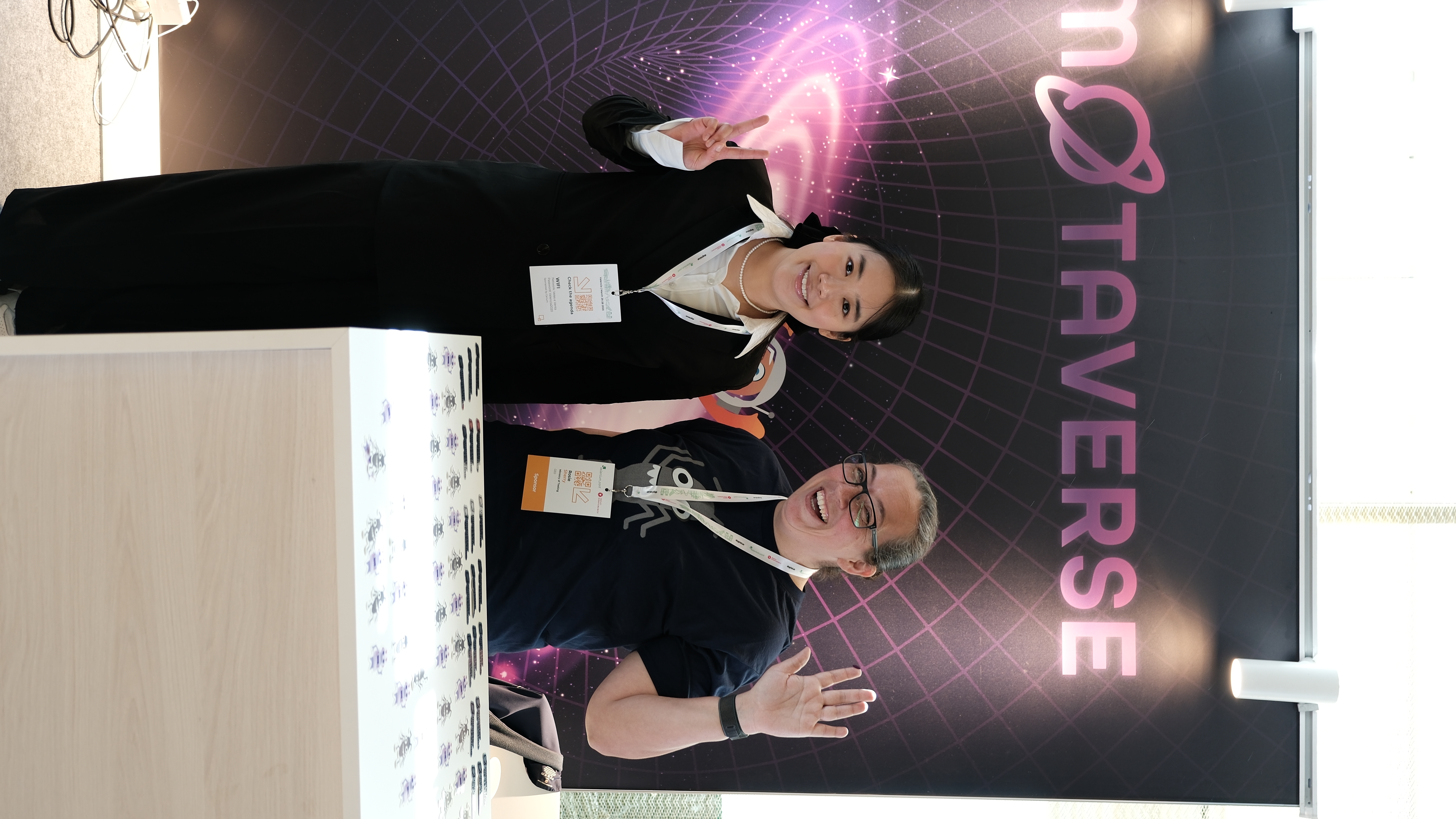 Amalie Shi, speaker at Selenium Conf 2025, stands smiling with Rosie Sherry at the Ministry of Testing stand, both posing in front of the MoTaverse backdrop with peace and wave gestures.