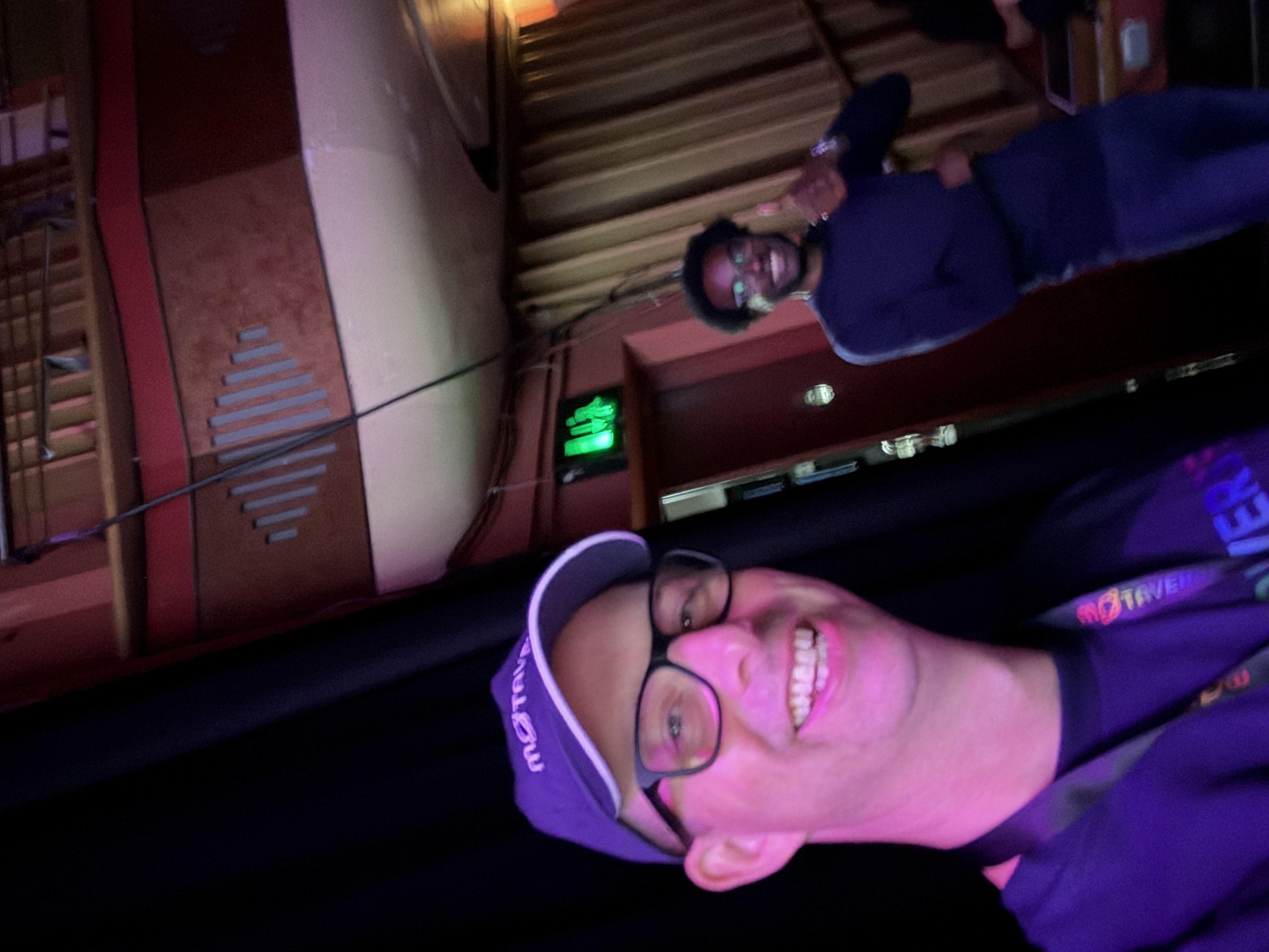 A smiling man takes a selfie, wearing a purple "MoTaverse" hat and lanyard, within a theatre space. His colleague sta... image