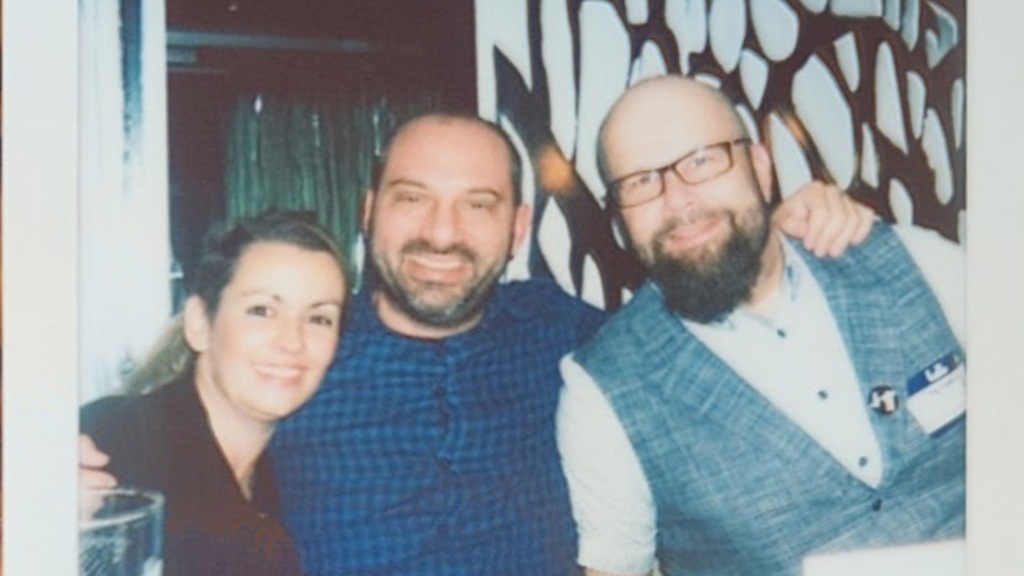 Diana, Petros, and Christian smiling together at a dinner table, with a decorative wall in the background.