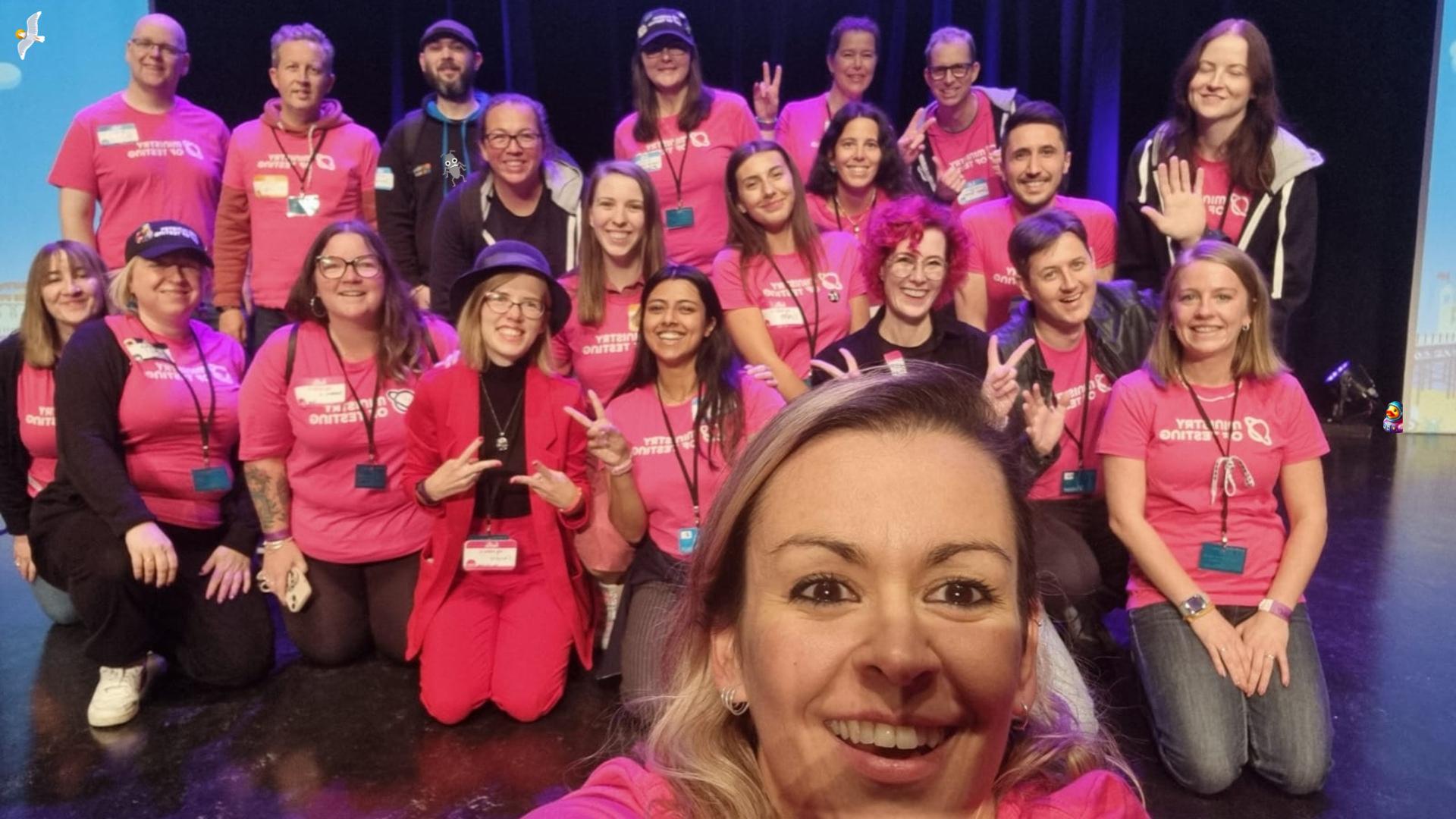 A medium sized group of people gathered on a stage smiling at the camera. Some are standing, some are kneeling. A woman is in the foreground holding the camera taking the selfie. Almost everyone is wearing a pink t-shirt with the ministry of testing logo on it. A seagull, bug and duck character have been added to the image.
