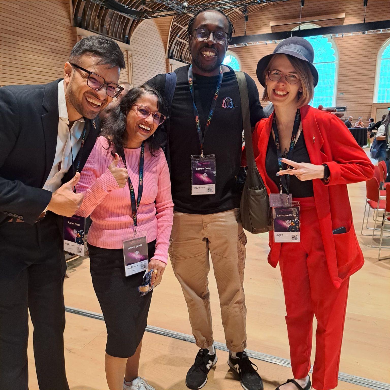 Four smiling teammates posing together at a conference hall, everyone wearing badges and casual-smart outfits, clearly happy to finally meet in person and capture the moment with a fun team photo.