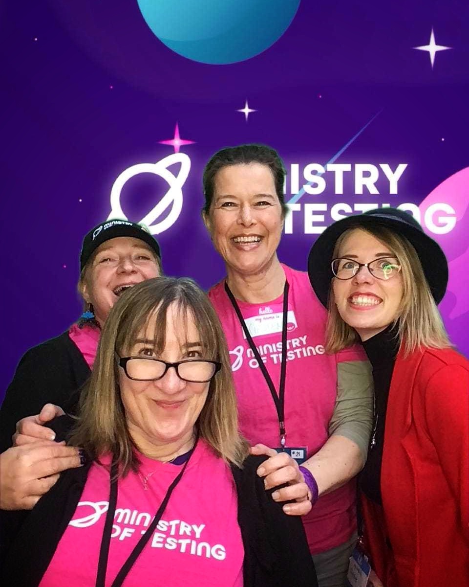 A group photo of four people at Ministry of Testing event Testbash 2024, standing in front of a purple backdrop with stars and planetary designs. Two people are wearing bright pink 'Ministry of Testing' t-shirts with lanyards, one person wears a black cap, and another person wears a blue hat and red jacket. All four are smiling broadly at the camera, and two are wearing glasses. The background features a space-themed design with a large blue planet and white stars.
