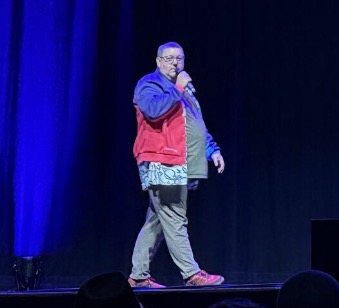 Ady, mike in hand, wearing a red and purple jacket, grey trousers and red trainers 