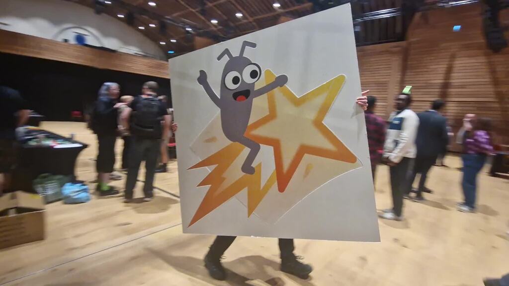 Someone carries a large board of the MoT bug mascot riding a shooting star around a wood-panelled conference hall at ... image
