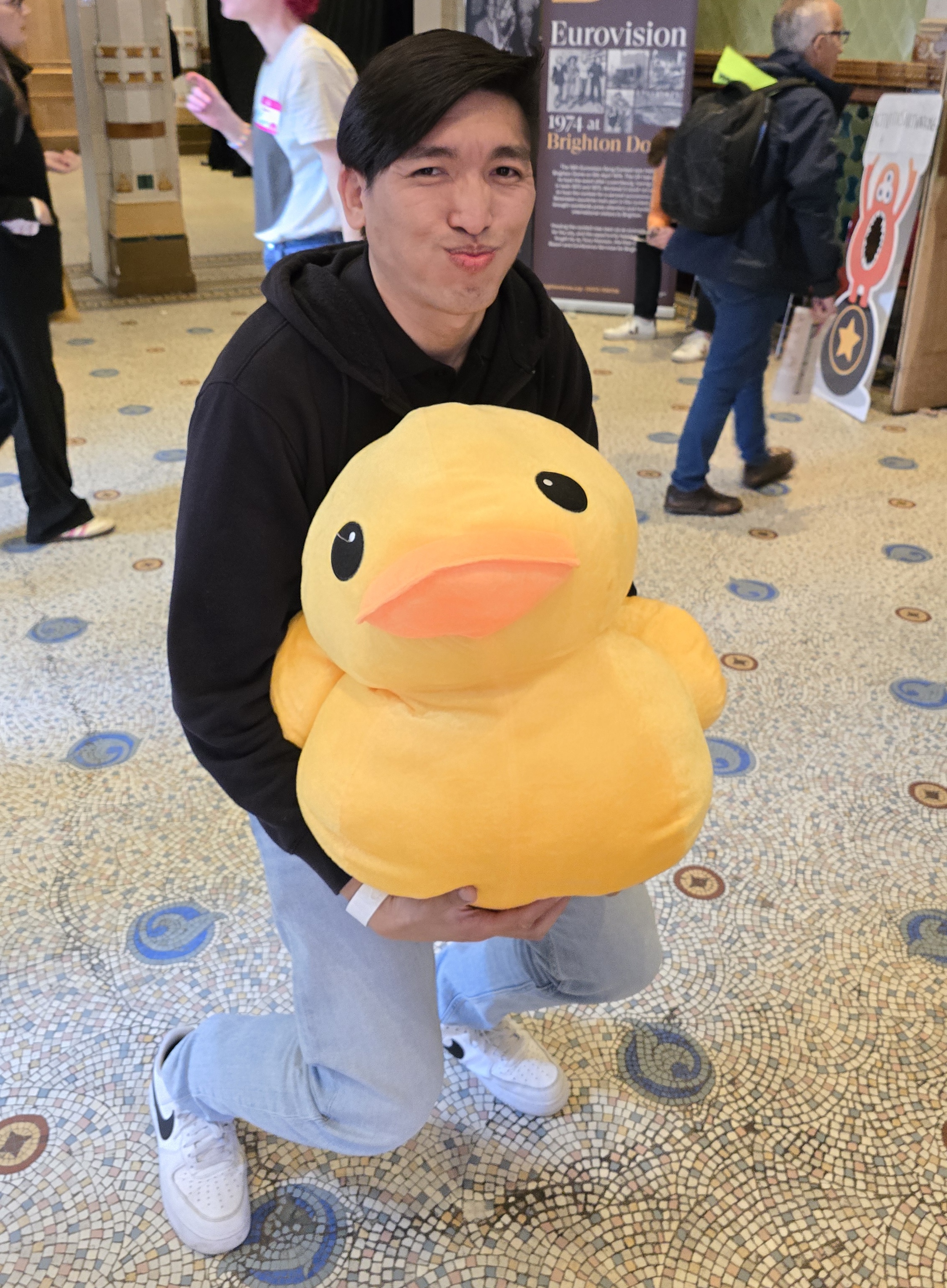 A man holds a large duck toy. Their knees are bent and their face looks concerned, suggesting that the duck is heavy. 