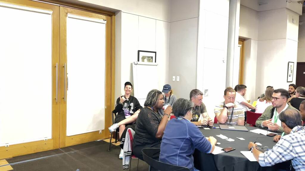 A slow pan across the Founder’s Room at the Brighton Dome, now filled with people. Every chair around the round tables is occupied, and participants are engaged in lively group discussions during a workshop session. The atmosphere is energetic and collaborative.
