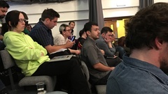 A group of people sat on auditorium-style chairs. At the end of the row is a man looking surprised.  image