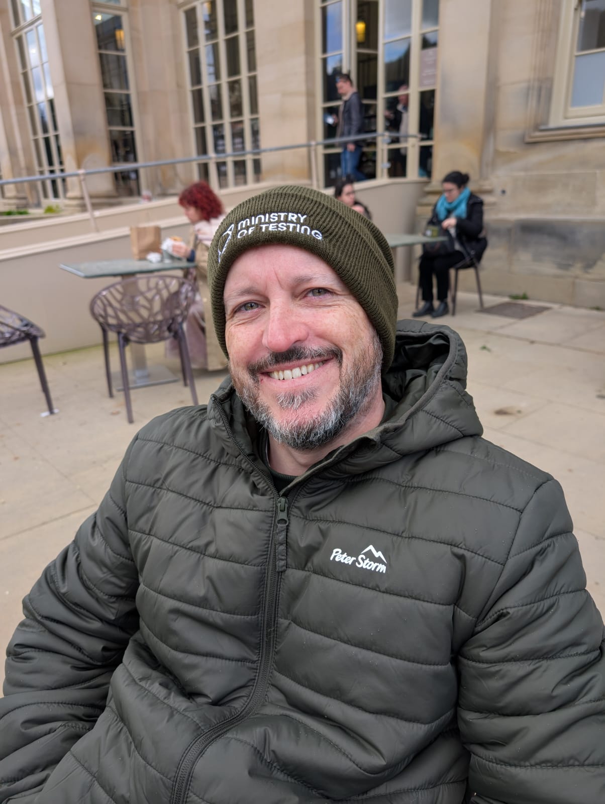 White bearded male sitting down smiling wearing a green ministry of testing beanie and a green coat image
