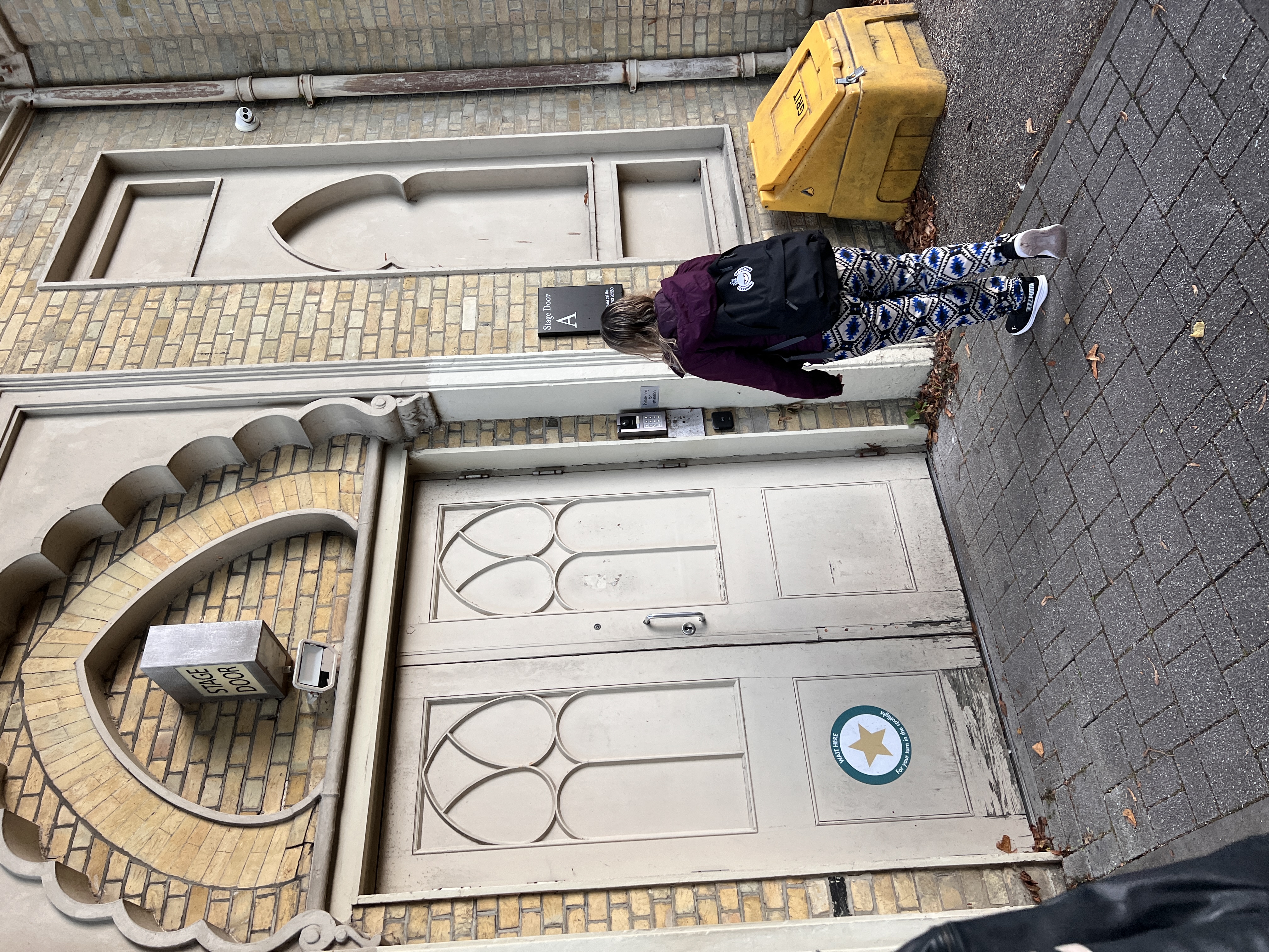 A woman with a rucksack stands outside a grand-looking door. There is a yellow container with the word GRIT on it. 