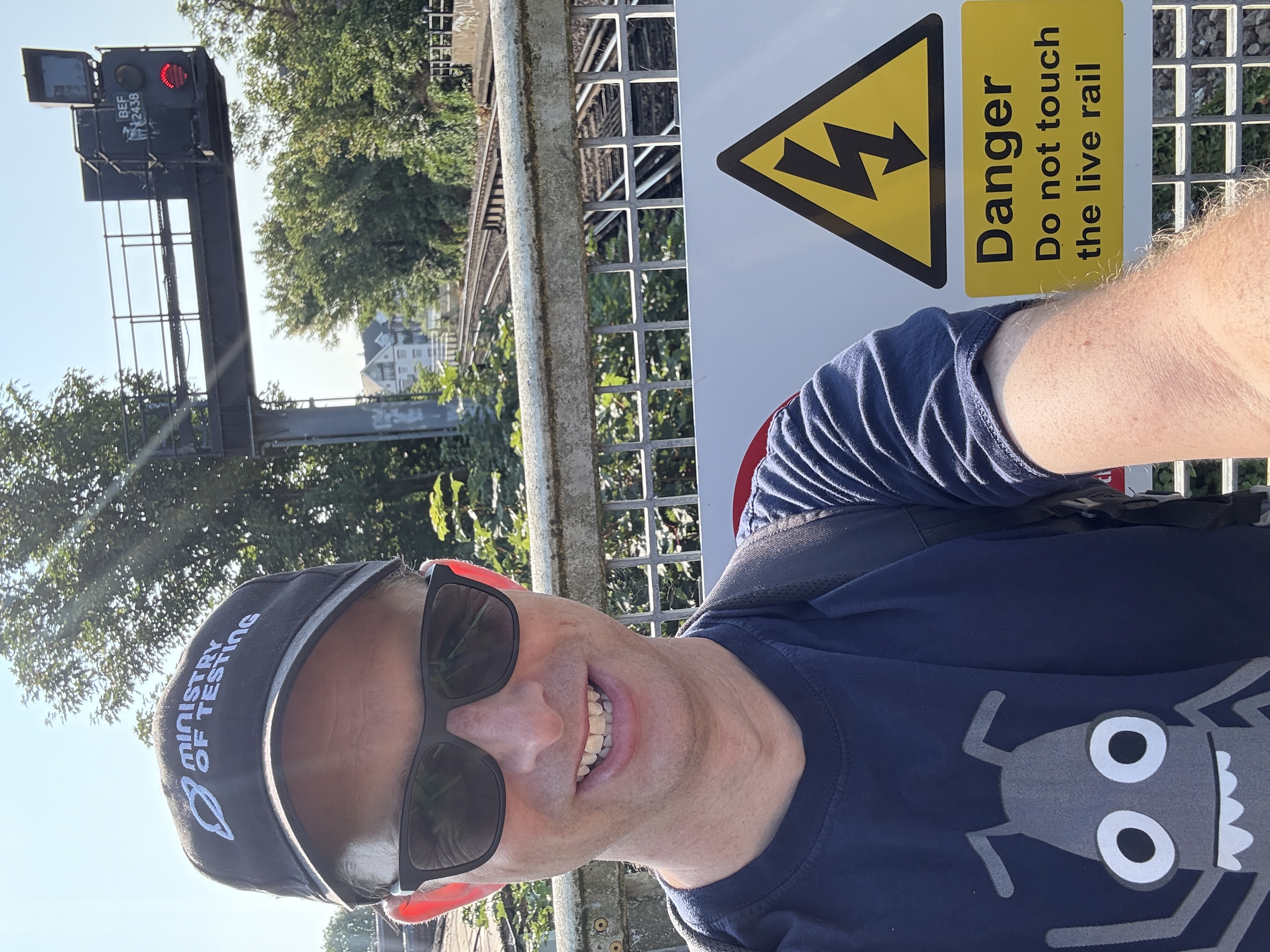 A man wearing a black cap and blue t-shirt with a big character on it stands at the end of a railway platform. 