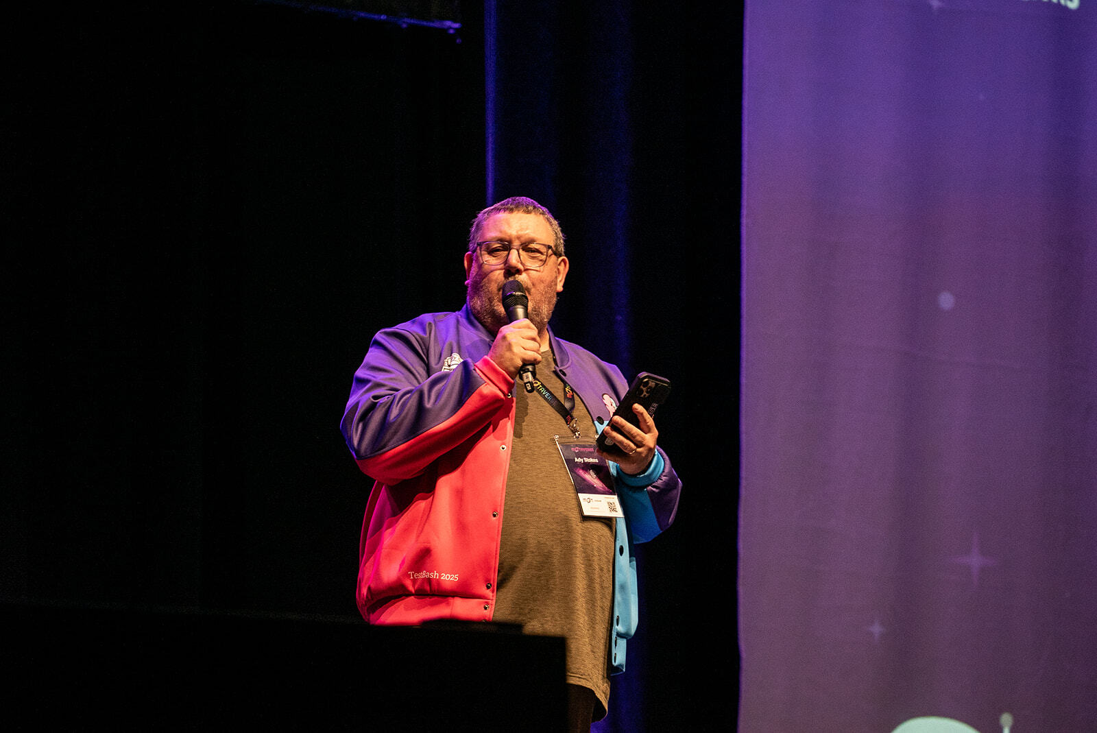 Ady, Mike in hand, on stage at TestBash / MoTaCon with a red and purple jacket, grey trousers and red trainers 