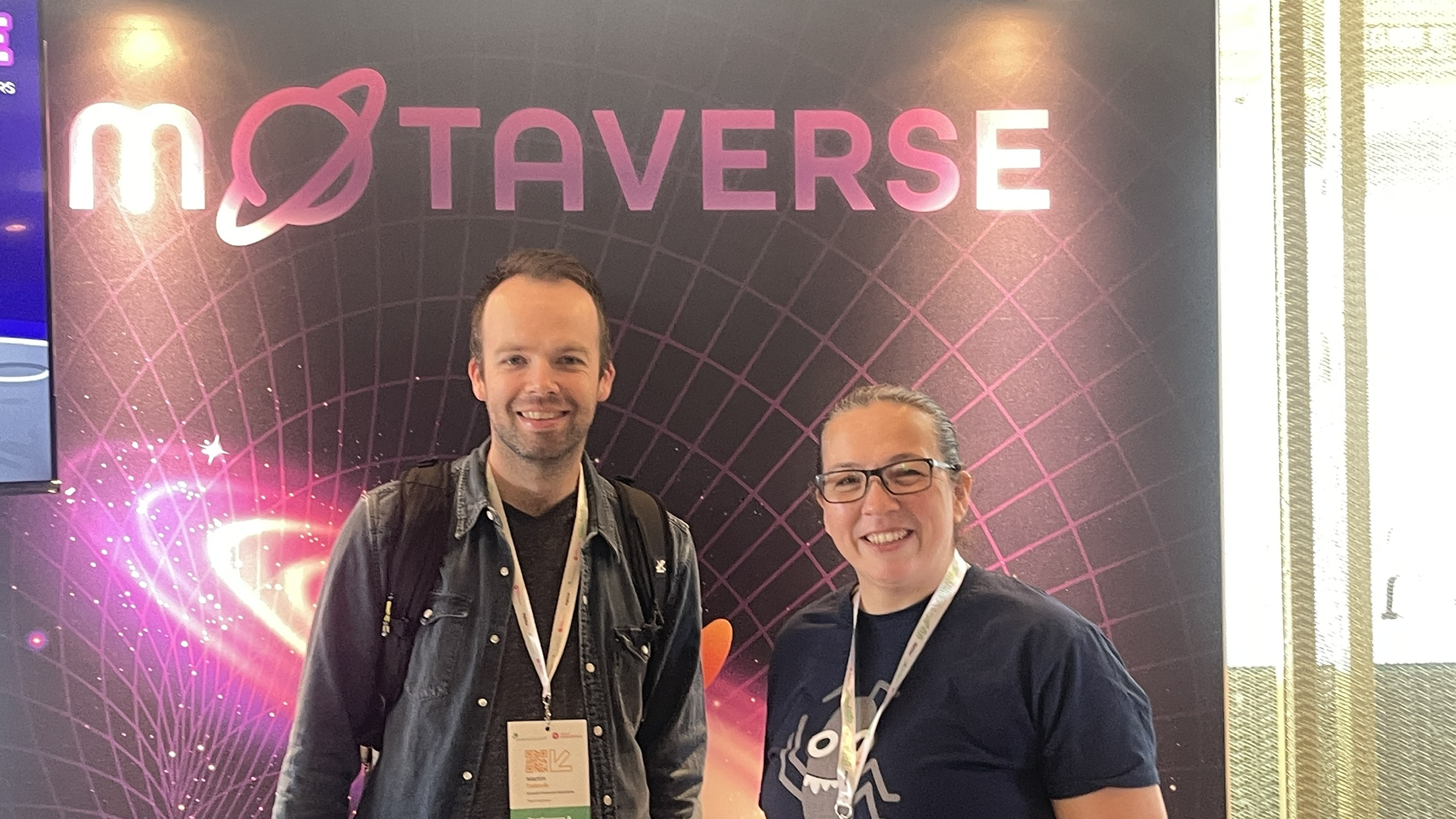 Rosie smiles with an attendee at the MoTaverse stand on Day 2 of SeleniumConf. The MoTaverse backdrop glows in the background, keeping the vibes cosmic.
