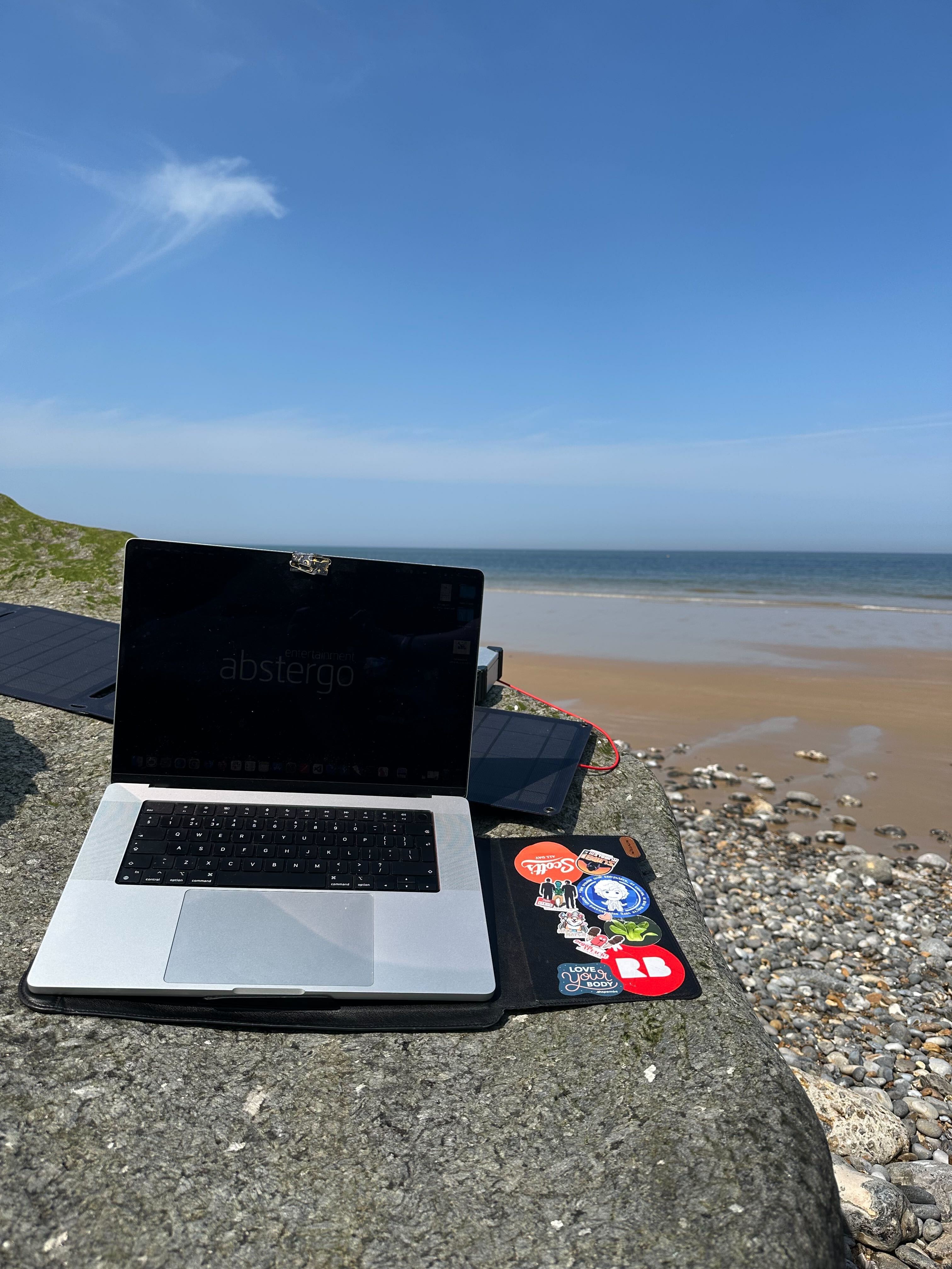 A silver MacBook with a black screen sits on a stone surface near a beach, facing the sea. The laptop's case is decorated with colourful stickers, including one that says "Love your body" and another with the Redbubble logo. Behind it, a portable solar panel is visible. The scene is bathed in bright sunlight under a clear blue sky, with calm waves and a sandy, pebbled shoreline in the background—suggesting remote work or study by the seaside.