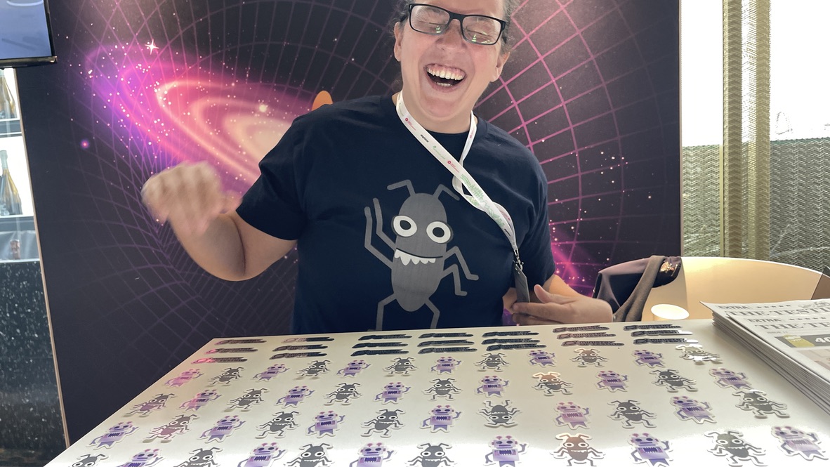 Rosie Sherry laughs joyfully at the Ministry of Testing stand during Selenium Conf 2025, wearing the popular bug t-shirt with a table full of MoT stickers in front of her. A stack of The Testing Planet newspapers is visible to the side.