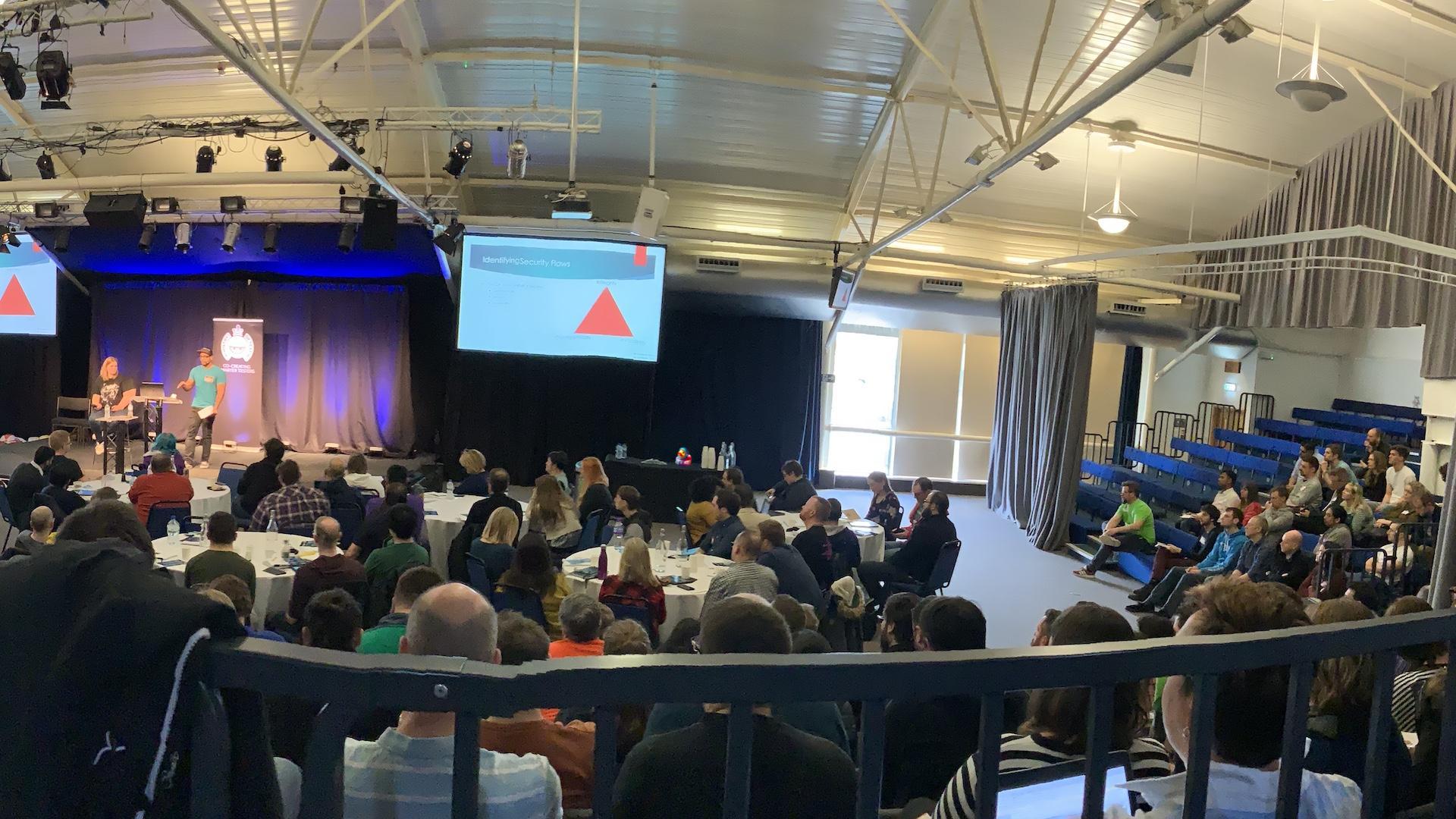 A conference auditorium space looking at the stage from the back of the room. Lots of people are listening to a talk. There is a woman and man on stage. A duck and bug character are hiding.