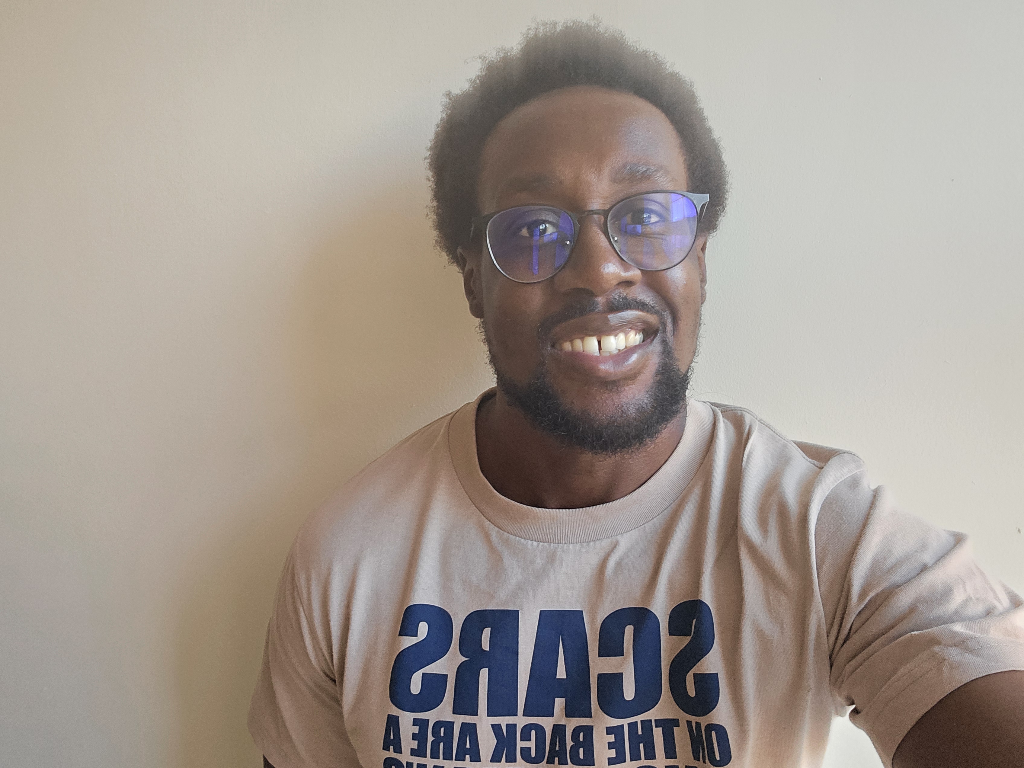 Selfie of Eamon Droko.

Eamon is a black man, with medium afro hair, wearing glasses with a blue tint.

He is smiling in the image.

He is wearing a beige coloured t-shirt that has dark blue text.