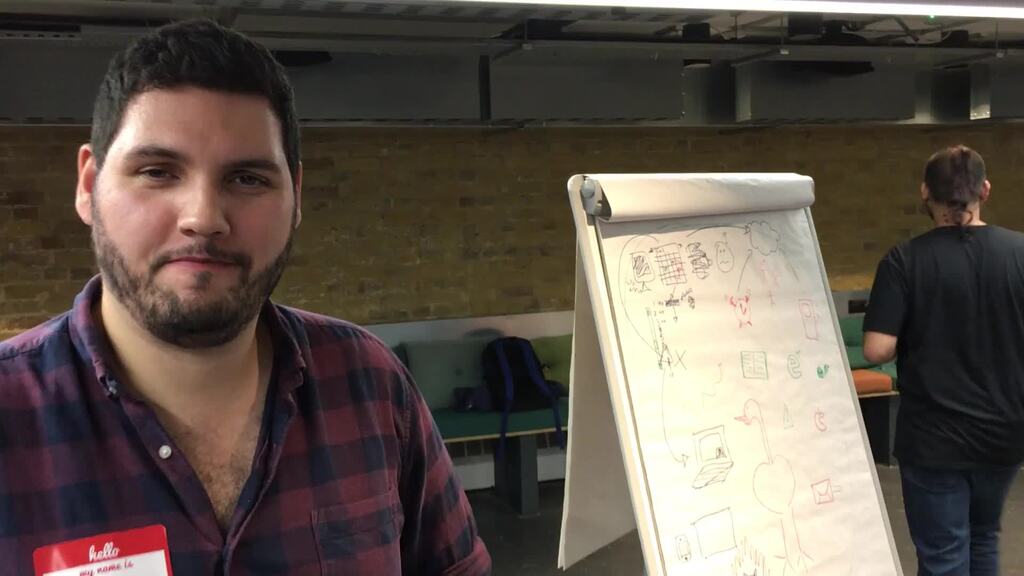 A man talking to camera with a flip-chart in the background.  image