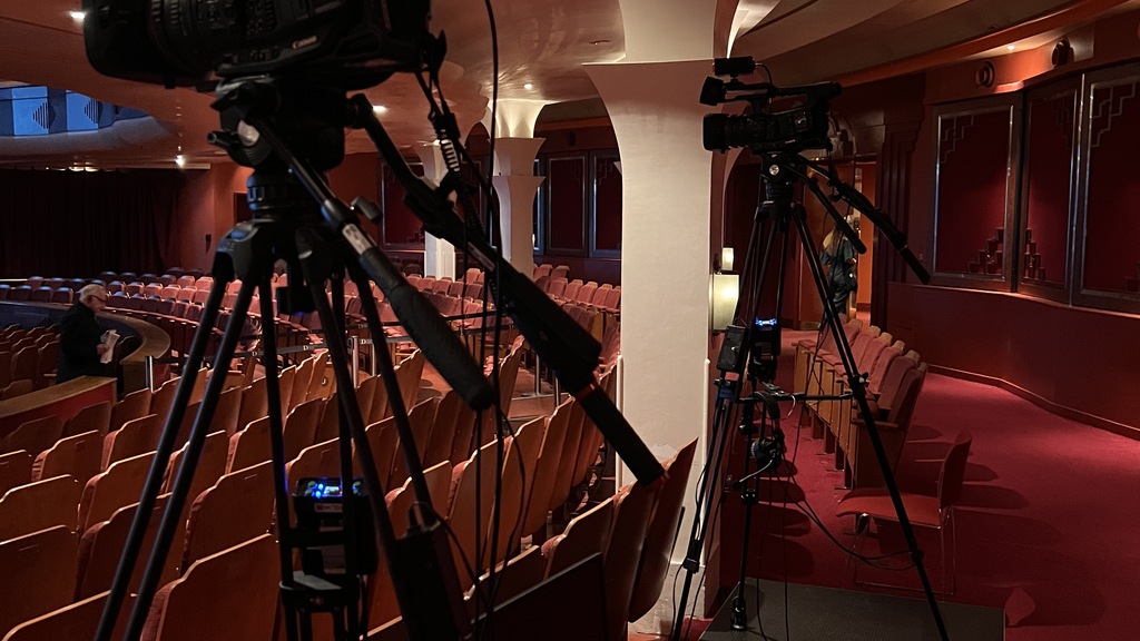 A photograph taken inside an empty theatre, looking towards the stage. Two video cameras on tripods are set up in the foreground. The theatre seats are red, and the walls are also a deep red colour, with ornate detailing and decorative columns. The lighting is dim, creating a warm, intimate atmosphere.