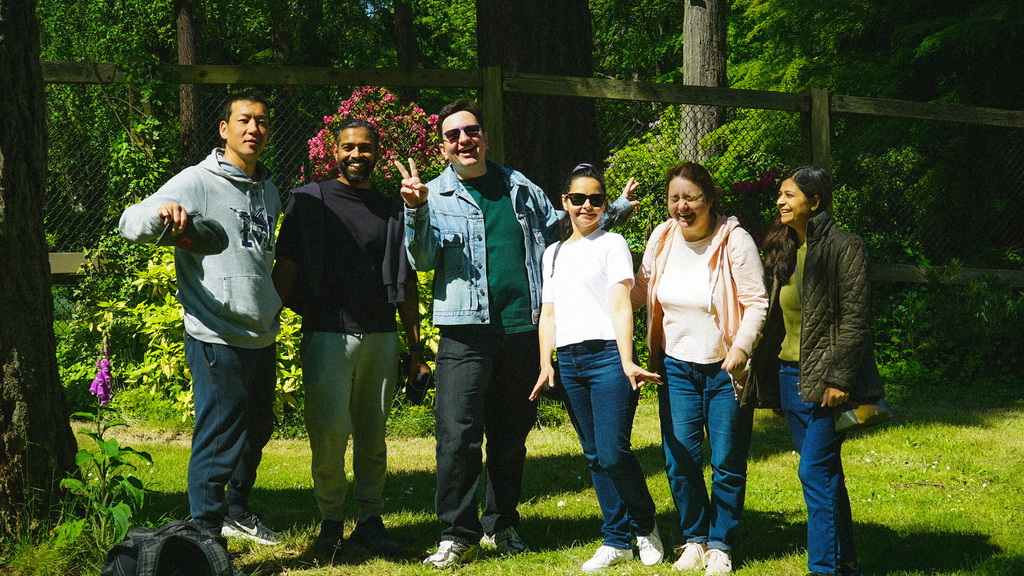 A group of six people smiling and posing together in a sunny, green park with tall trees and flowering bushes behind them. Everyone looks relaxed and cheerful, with one person flashing a peace sign.