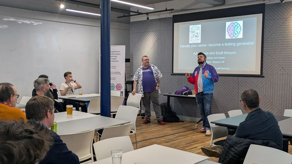 Ady and Scott stand in front of a screen with the session title how to elevate your career; become a testing generalist. Scott is wearing a pink and blue jacket with purple shoulders and Ady has a MoT planet logo Hawaiian style shirt. A group sat at several tables are looking at them. 