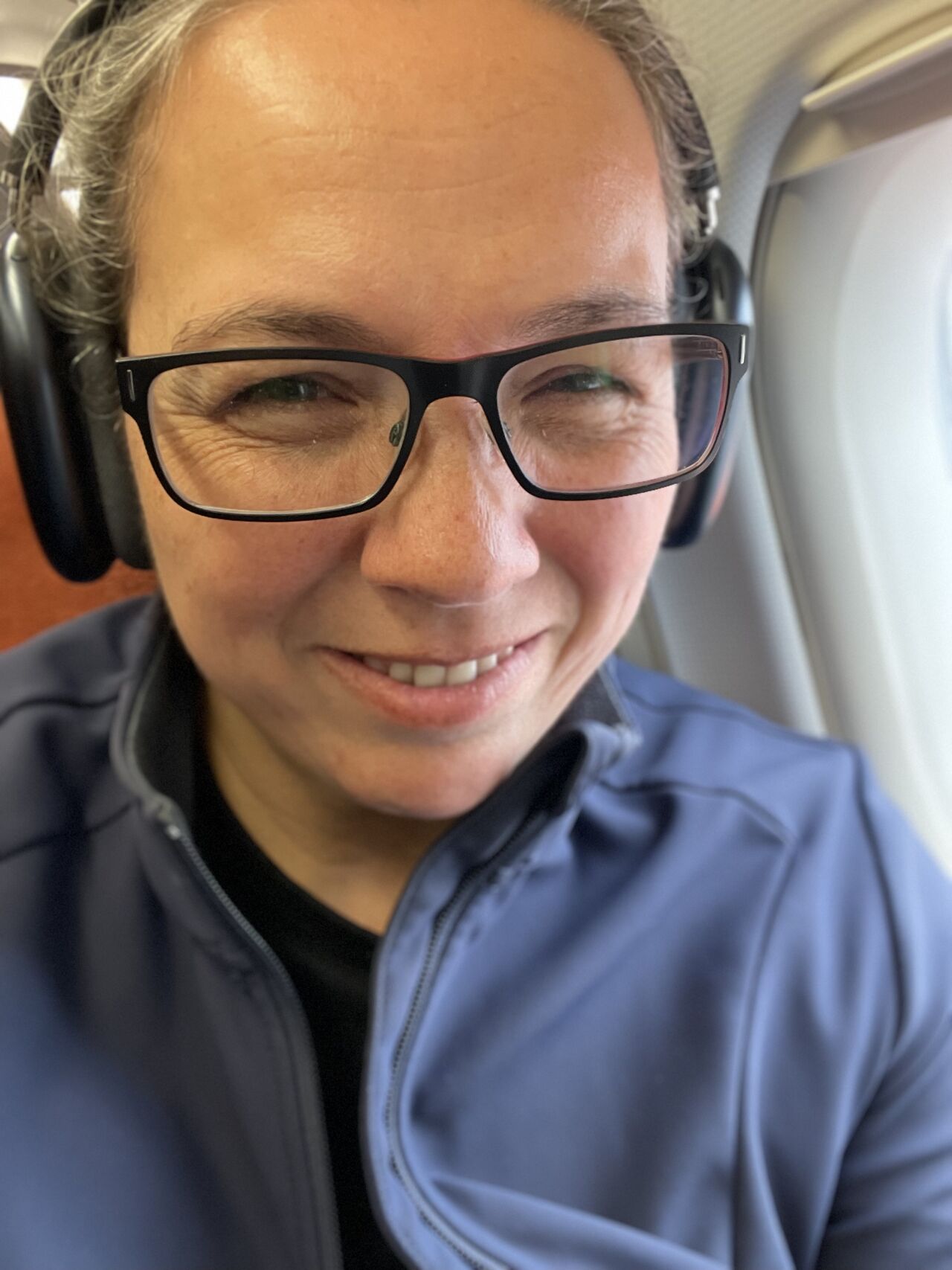 A selfie of Rosie on an aeroplane. She has headphones on, dark rimmed glasses and a blue jacket.