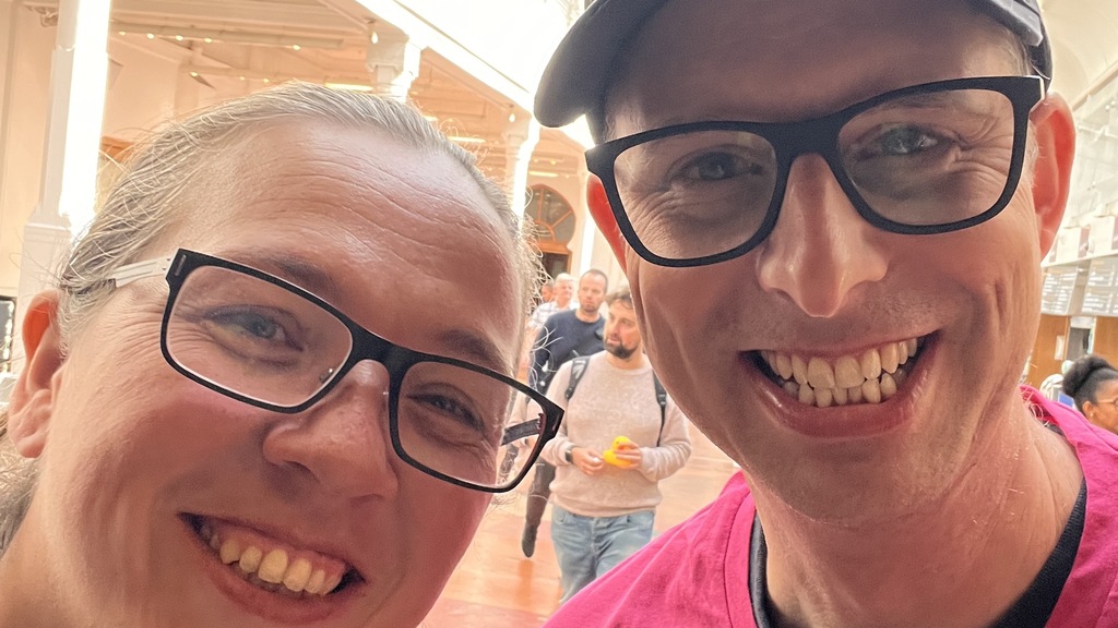 A smiling man and woman take a selfie indoors, with a crowd of people in the blurred background. The man wears a black baseball cap and a pink shirt, both with the "Ministry of Testing" logo.