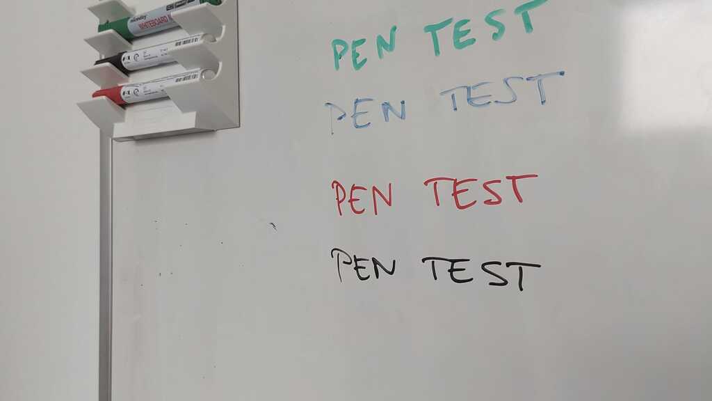 Whiteboard with four “PEN TEST” texts in green, blue, red, and black. The blue text is faint, hinting it’s a weak marker. A wall-mounted holder shows only green, red, and black pens - blue is missing.