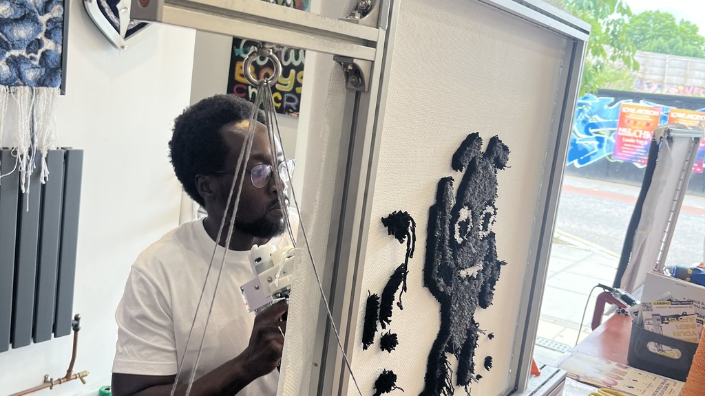 A picture of Eamon Droko at Cheeky studio who is rug tufting a rug of Bug using the classic grey, black and white colours. He is focused and concentrating whilst using the rug tufting gun. 