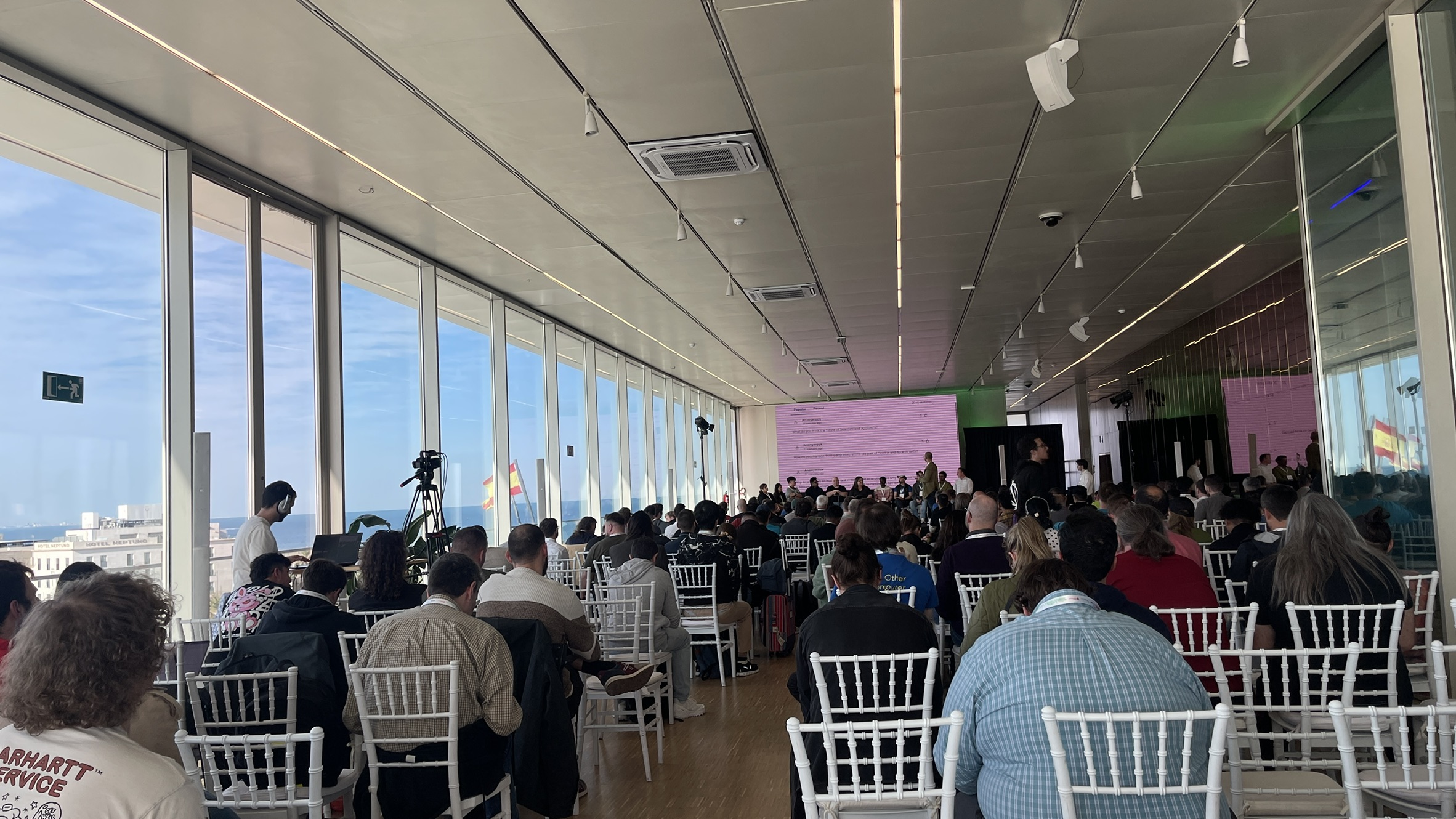 A wide-angle shot of the main conference room at SeleniumConf 2025. Rows of attendees are seated facing a stage and large screen, with a wall of glass windows revealing a bright blue sky and Mediterranean Sea beyond.