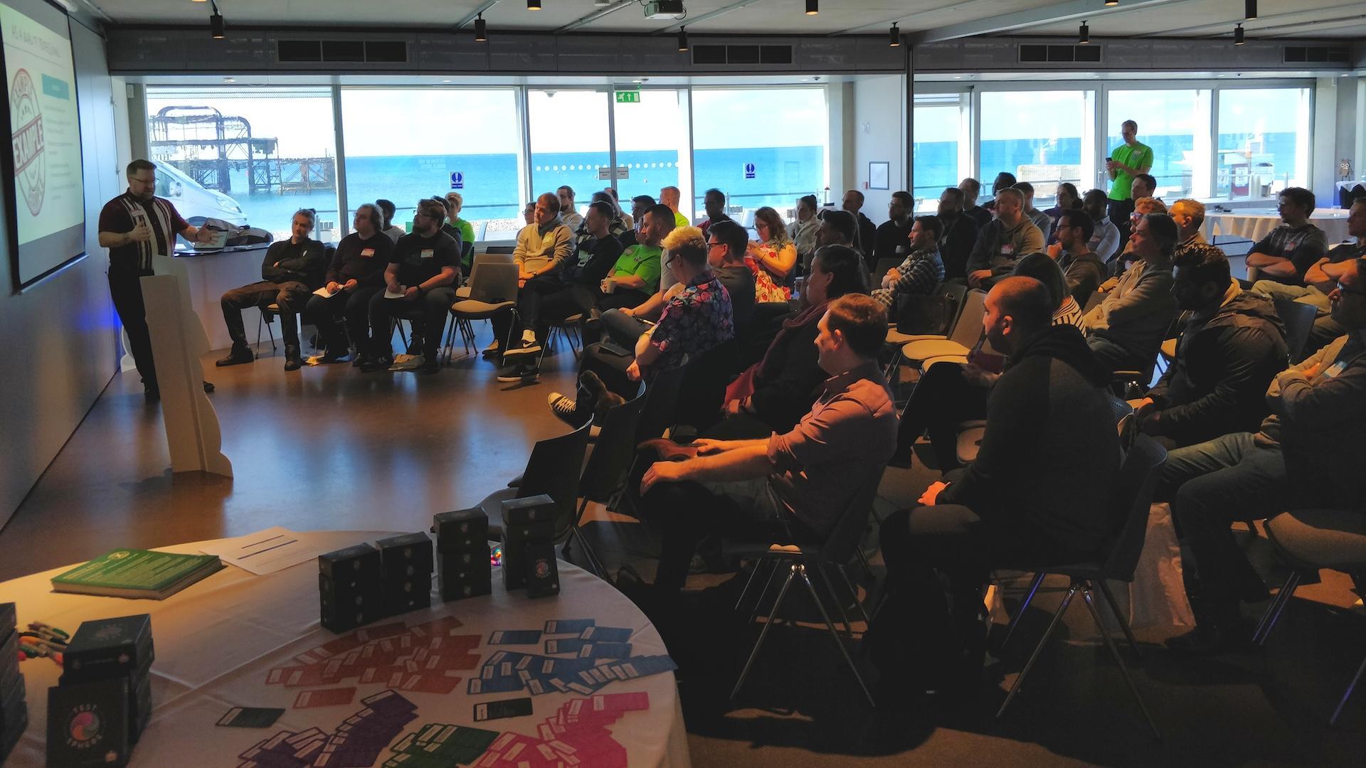 A medium size event space overlooking the sea. A group of people are watching a presentation. There is a bug and duck character hiding.