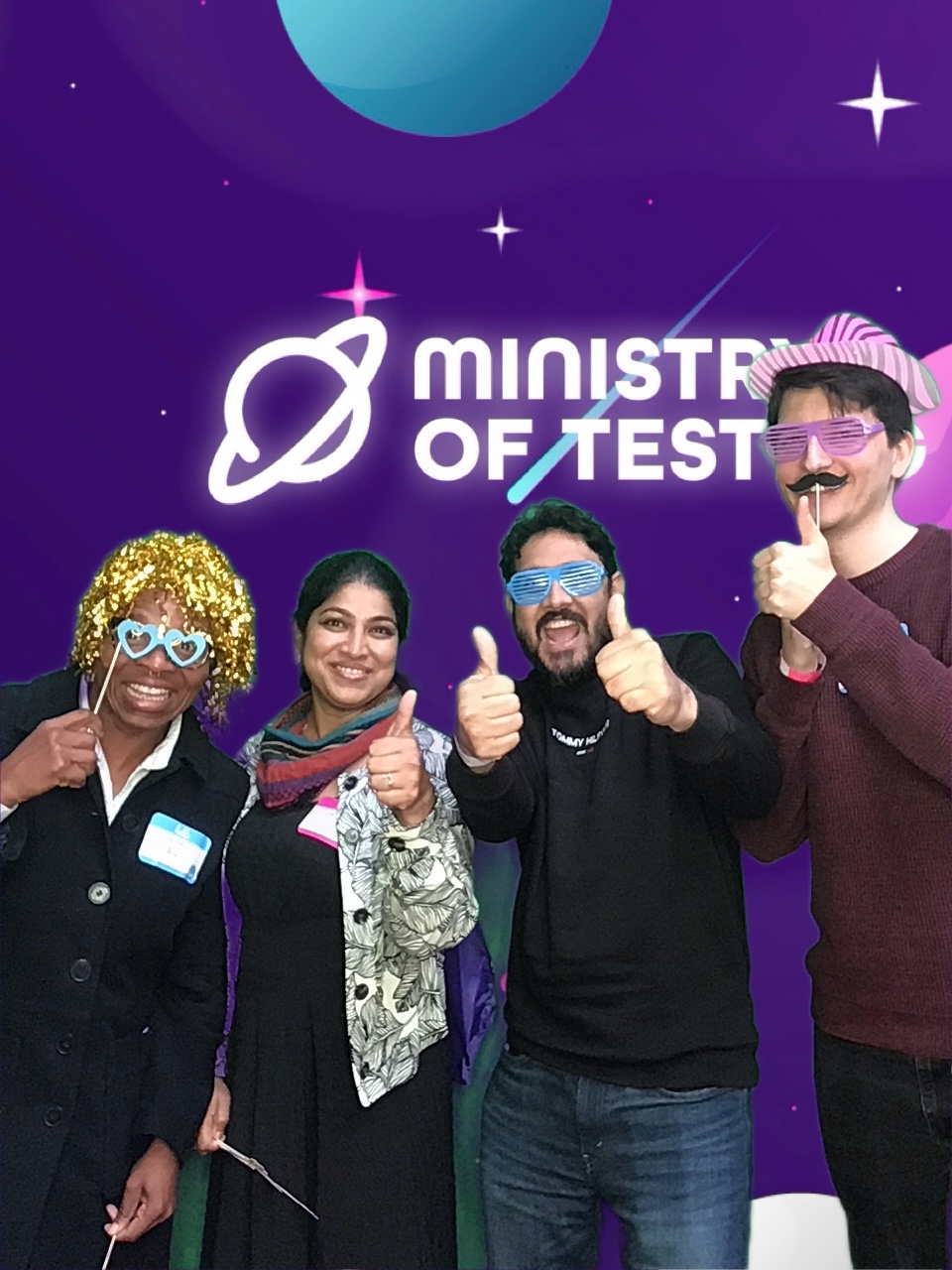 Here we are getting thumbs up from 3/4 TestBash 2024 attendees in the photobooth. We have a mixture of silly accessories making an appearance in this photo. From a gold tinsel wig, to glasses on a stick, to pink zebra print hats, moustaches on a stick and colourful pretend glasses. This picture is full of energy and excellent vibes, and we see that from the expressions and gestures given at this moment. They are also kind of in heigh order too from left to right, whether or not they meant to be, it looks pretty good in the picture.

Background: The green screen backdrop depicts a simple illustrated outer space scene. The main colour is purple, with cute stars and planets in shades of pink, blue, and white. About a third of the way down the screen, a logo appears. It reads "Ministry of Testing" in bold white text with a blue shooting star behind it. Next to the text, in the same bold white style, is a line drawing of the planet Saturn.

