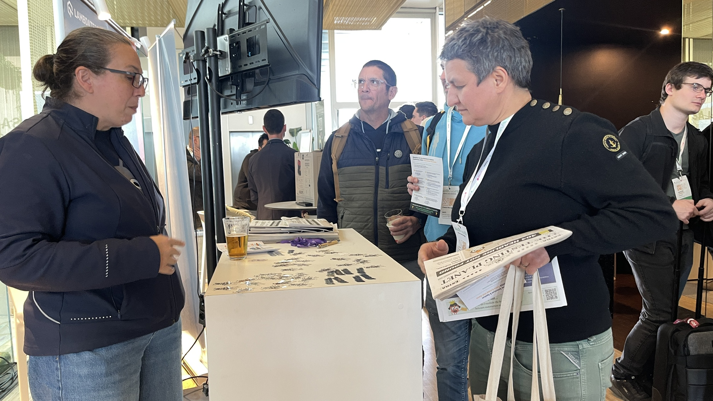 Rosie chats with conference attendees at the Ministry of Testing stand during the final rush to complete the sponsor quiz for the raffle entry. The stand is buzzing, the table is scattered with bug stickers and Testing Planet newspapers, and attendees are clutching raffle cards and drinks.