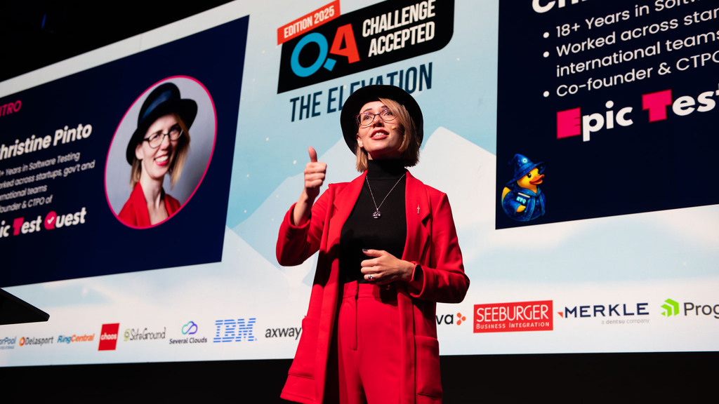 Christine Pinto speaking on stage at the QA: Challenge Accepted 2025 conference in Sofia, Bulgaria. She’s wearing a red suit and black hat, giving a thumbs-up while presenting Epic Test Quest, with her intro slide displayed behind her.