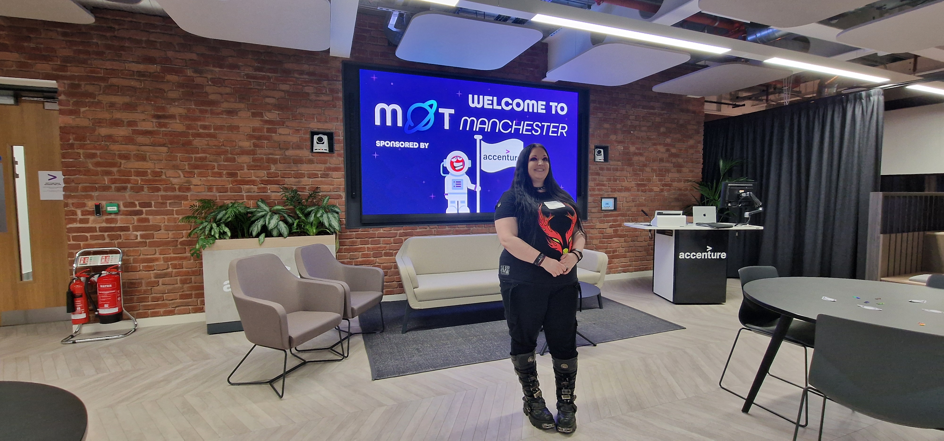 A person with long dark hair stands smiling in a modern office lounge, wearing a black t-shirt with a red graphic, bl... image