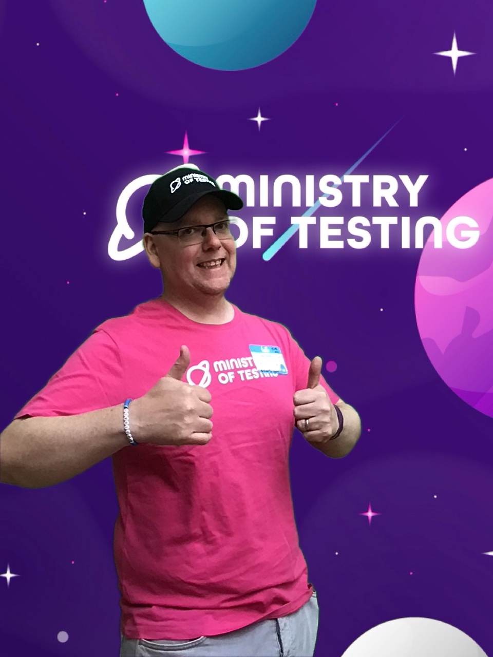 This attendee wears a black ministry of testing cap, with a pink ministry of testing tshirt and holds 2 thumbs up. They wear glasses and have a smile on their face. 

Background: The green screen backdrop depicts a simple illustrated outer space scene. The main colour is purple, with cute stars and planets in shades of pink, blue, and white. About a third of the way down the screen, a logo appears. It reads "Ministry of Testing" in bold white text with a blue shooting star behind it. Next to the text, in the same bold white style, is a line drawing of the planet Saturn.