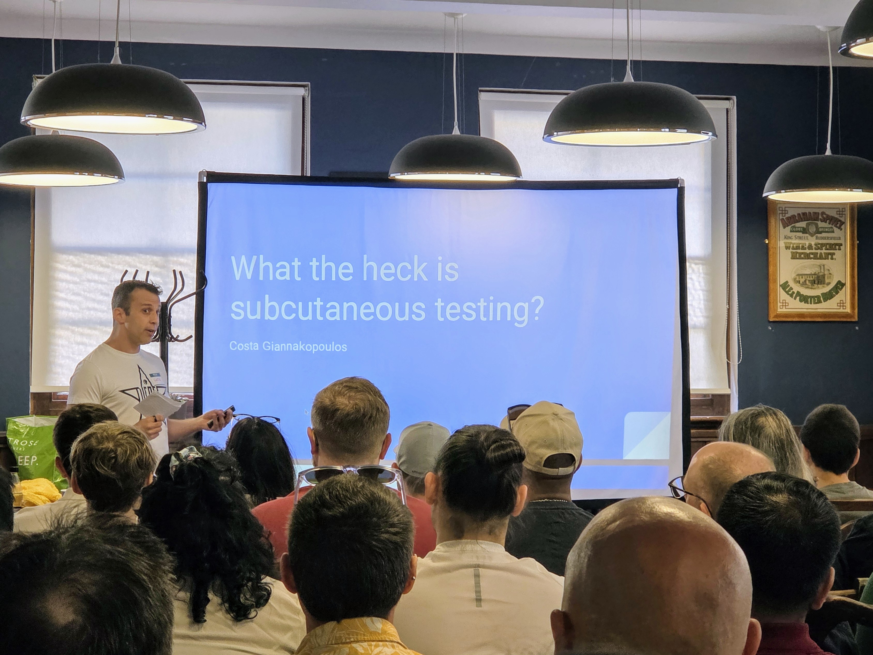Costa Giannakopoulos speaking to a full room at Leeds Testing Atelier, with a slide behind him reading “What the heck is subcutaneous testing?”