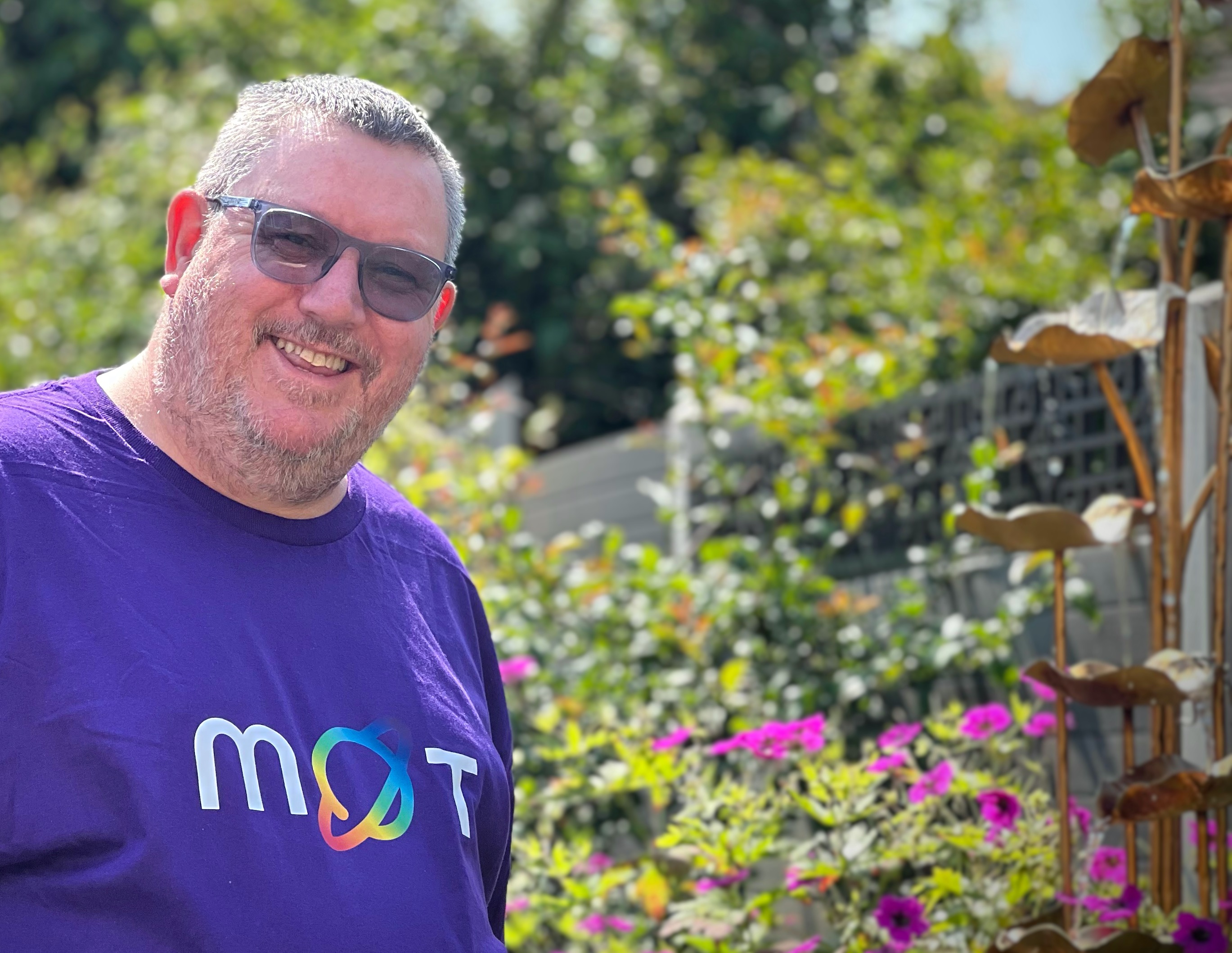 Ady is in the garden wearing dark glasses and a purple t-shirt with a rainbow M O T graphic. The O is the planet logo. 