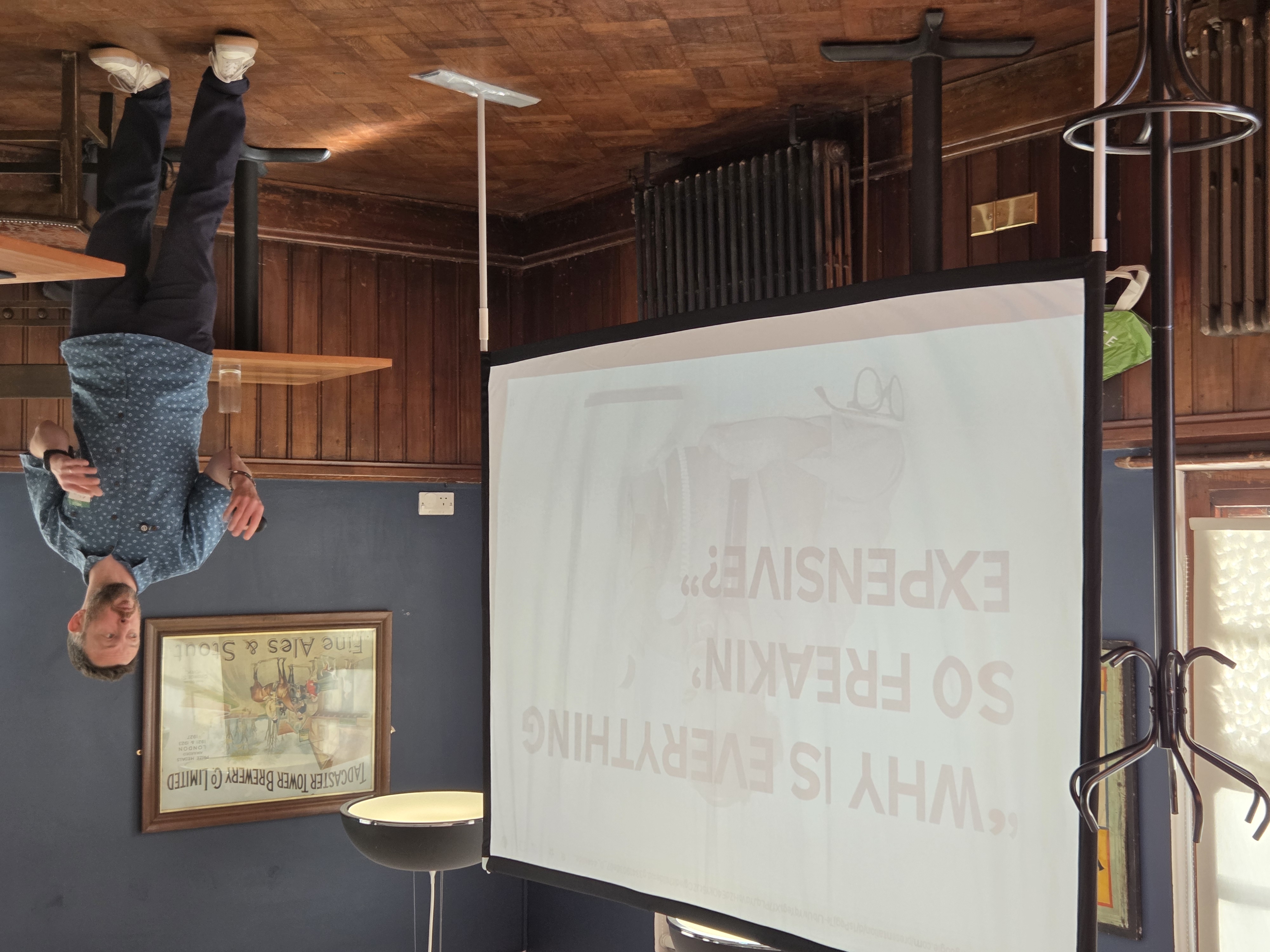 Clement Pickering speaking at Leeds Testing Atelier next to a large slide that reads, “Why is everything so freakin’ expensive?”