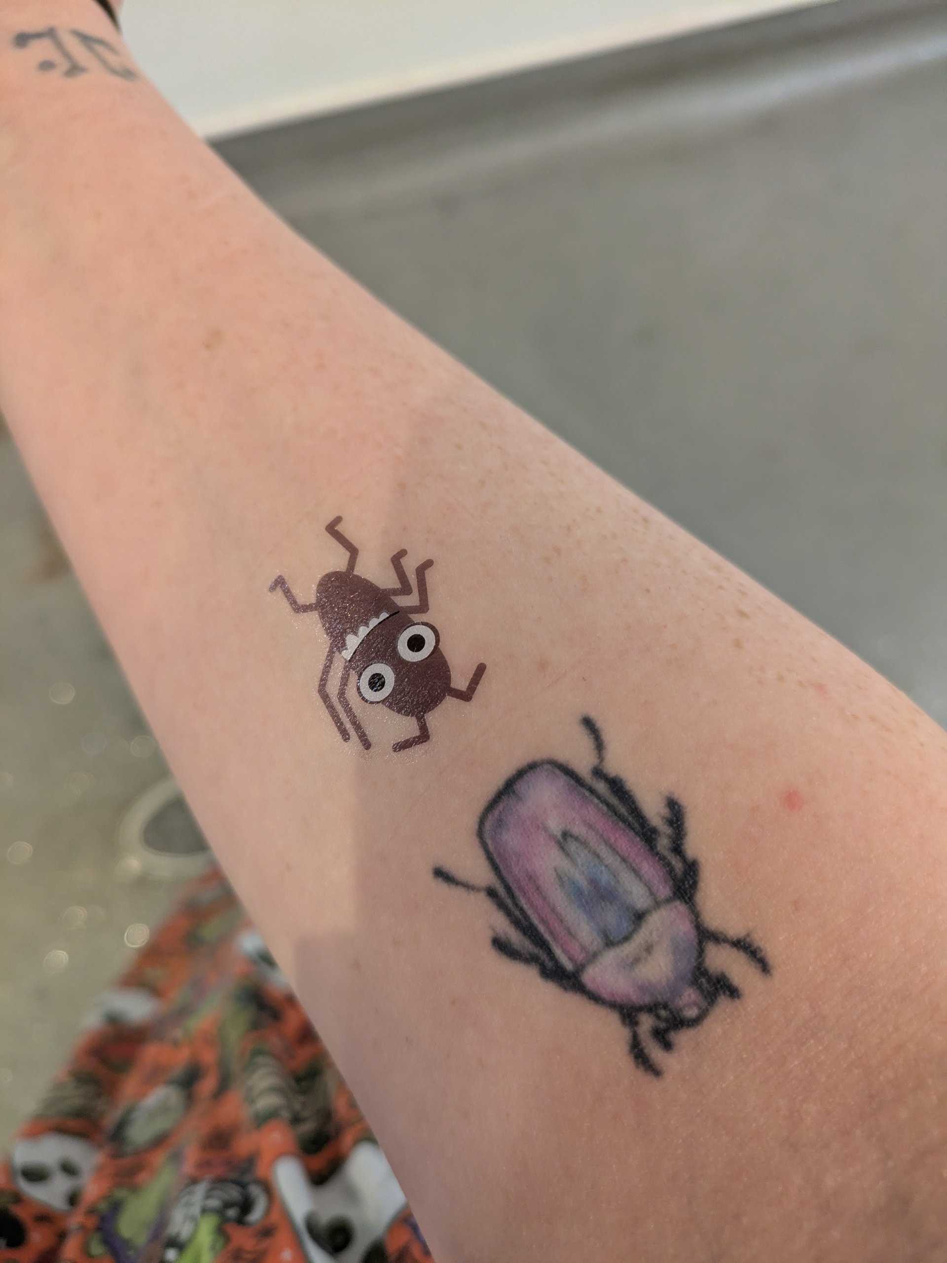 photo of a right arm with two bug tattoos: One grey and temporary one purple.
There is also a rorshach symbol on the right hand side of the wrist.
An orange dress with green frogs in various halloween costumes can also be seen.