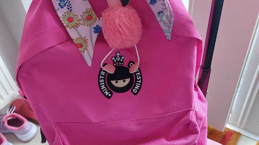 A pink suitcase with a pink "Ministry of Testing" themed rucksack sitting on top of it. The rucksack has been decorat... image