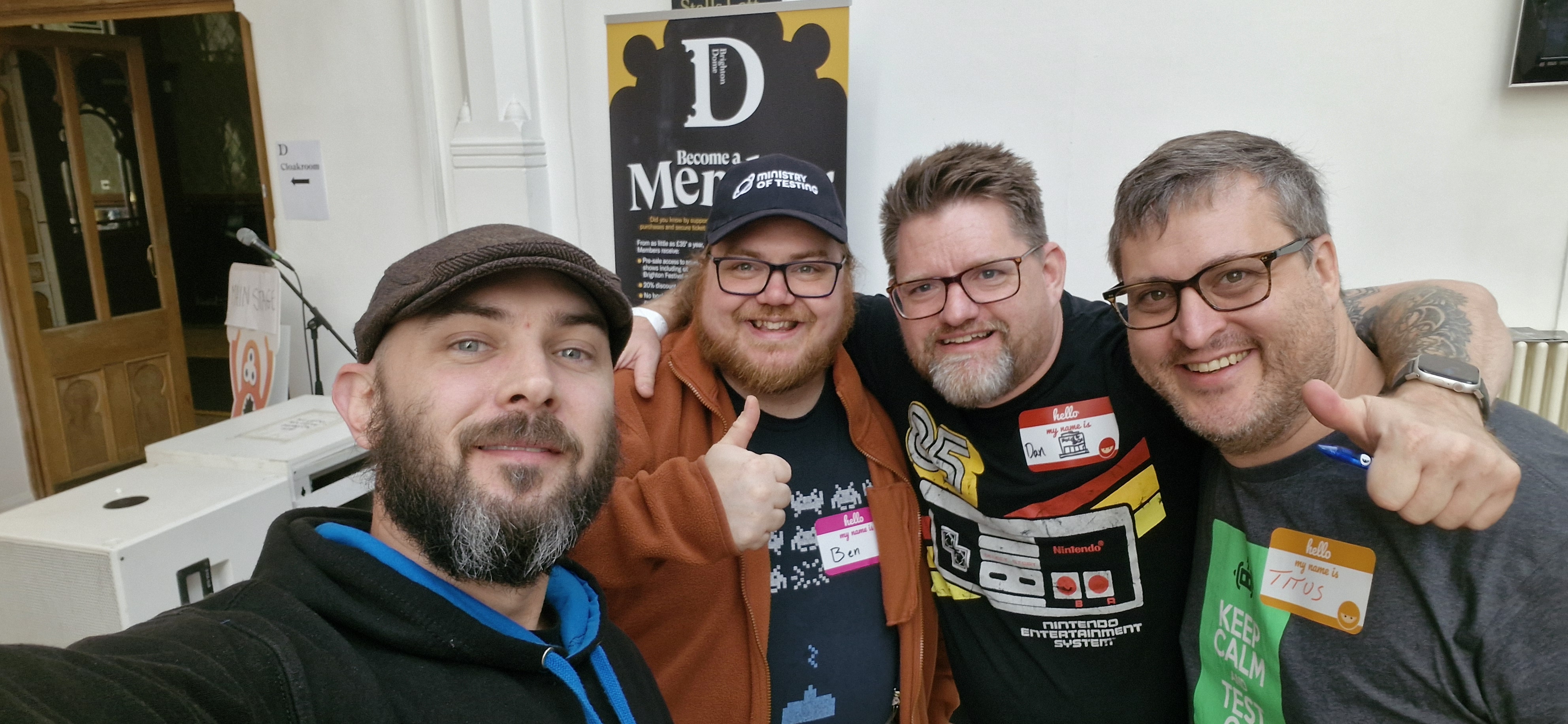 Four men are smiling and posing for a selfie indoors. One is wearing a cap, others have name tags. They all appear cheerful and are standing closely together.